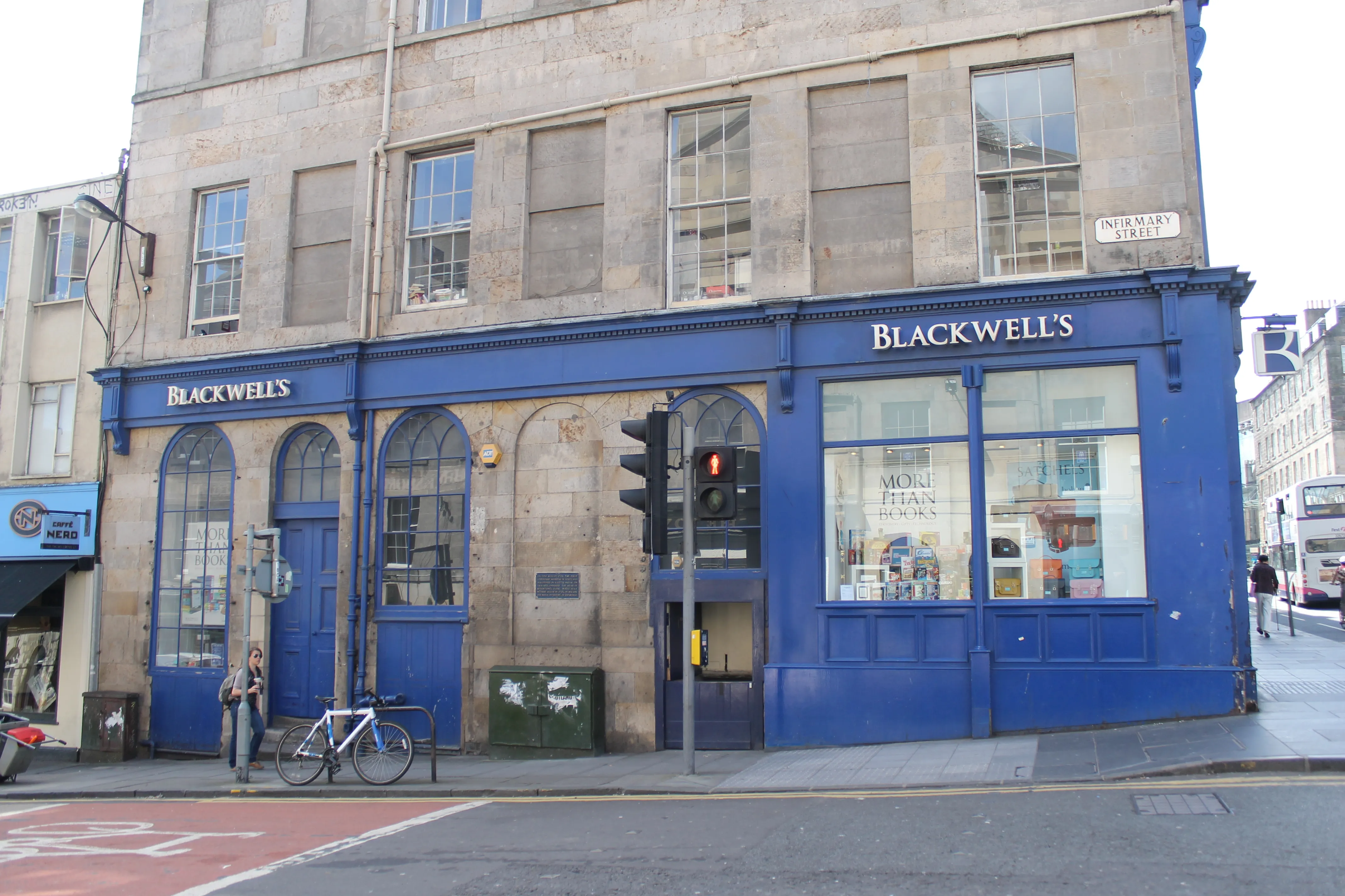 Widok z poziomu ulicy na zewnętrzną stronę księgarni Blackwell's Bookshop, ukazujący wyrazistą niebieską fasadę z szyldem BLACKWELL'S, łukowate okna z wystawami książek, sygnalizację świetlną oraz rower zaparkowany przed lokalem przy Broad Street.