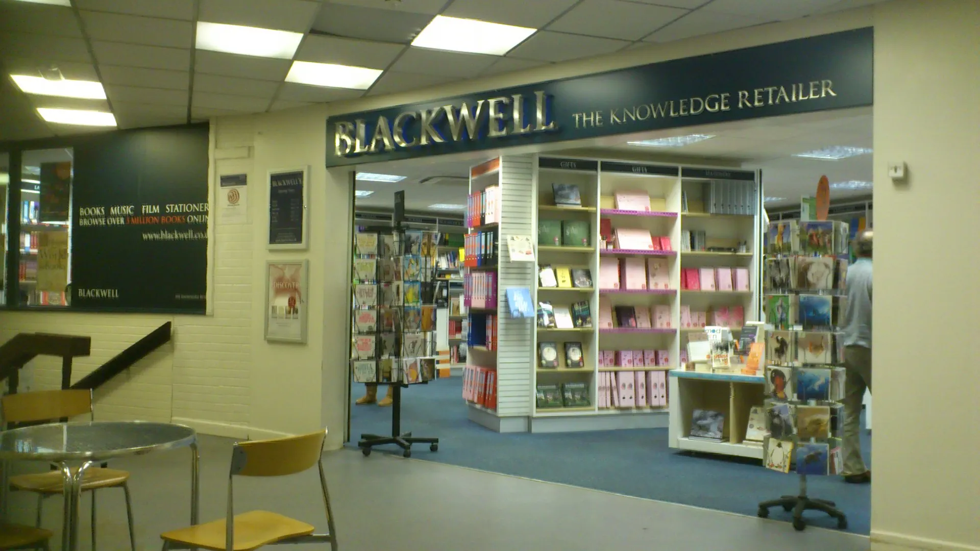 Wnętrze księgarni Blackwell's z widocznym szyldem 'Blackwell The Knowledge Retailer' nad wejściem do działu z książkami, z regałami, ekspozycjami i miejscami do siedzenia na pierwszym planie