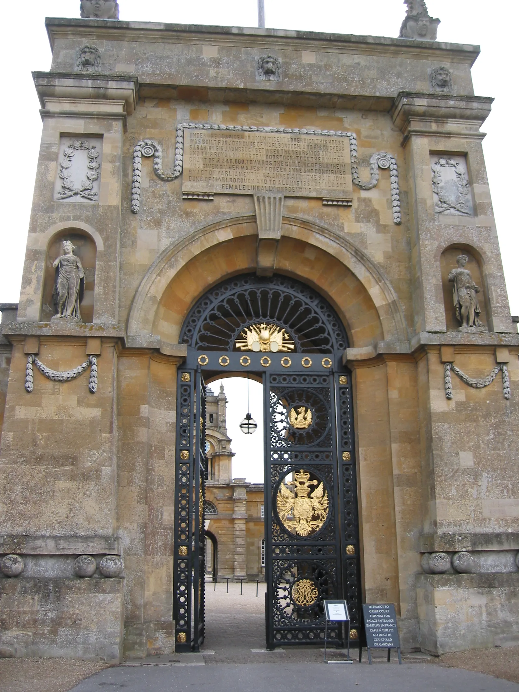Ozdobne czarno-złote żelazne bramy umieszczone w dużym kamiennym łuku przy wejściu do Blenheim Palace, flankowane przez klasyczne posągi w niszach.