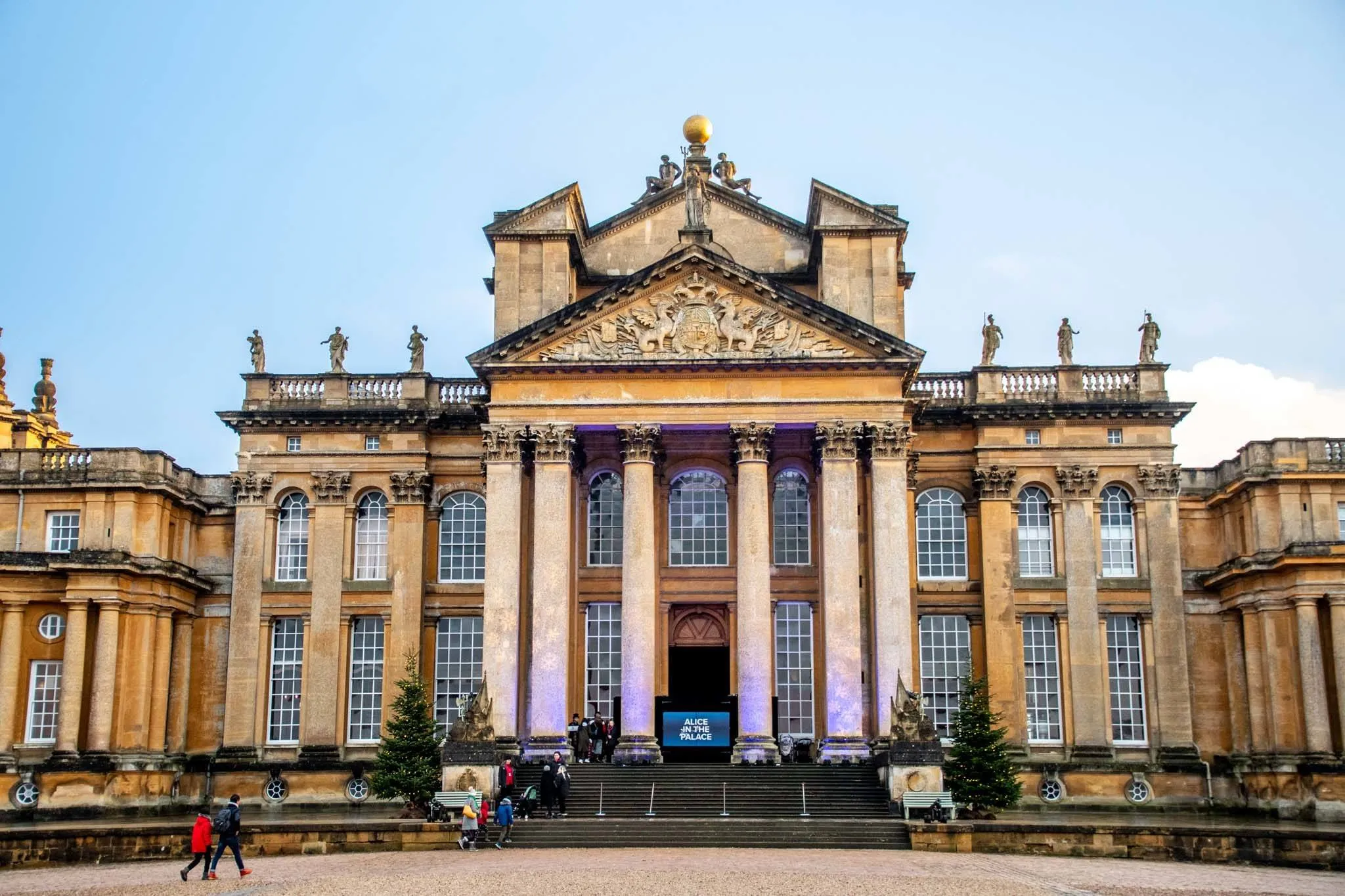 Pełny widok z przodu barokowej fasady Blenheim Palace z wysokimi kolumnami korynckimi, trójkątnym frontonem z płaskorzeźbami, złotą kulą na dachu oraz szerokimi schodami prowadzącymi do głównego wejścia pod błękitnym niebem.