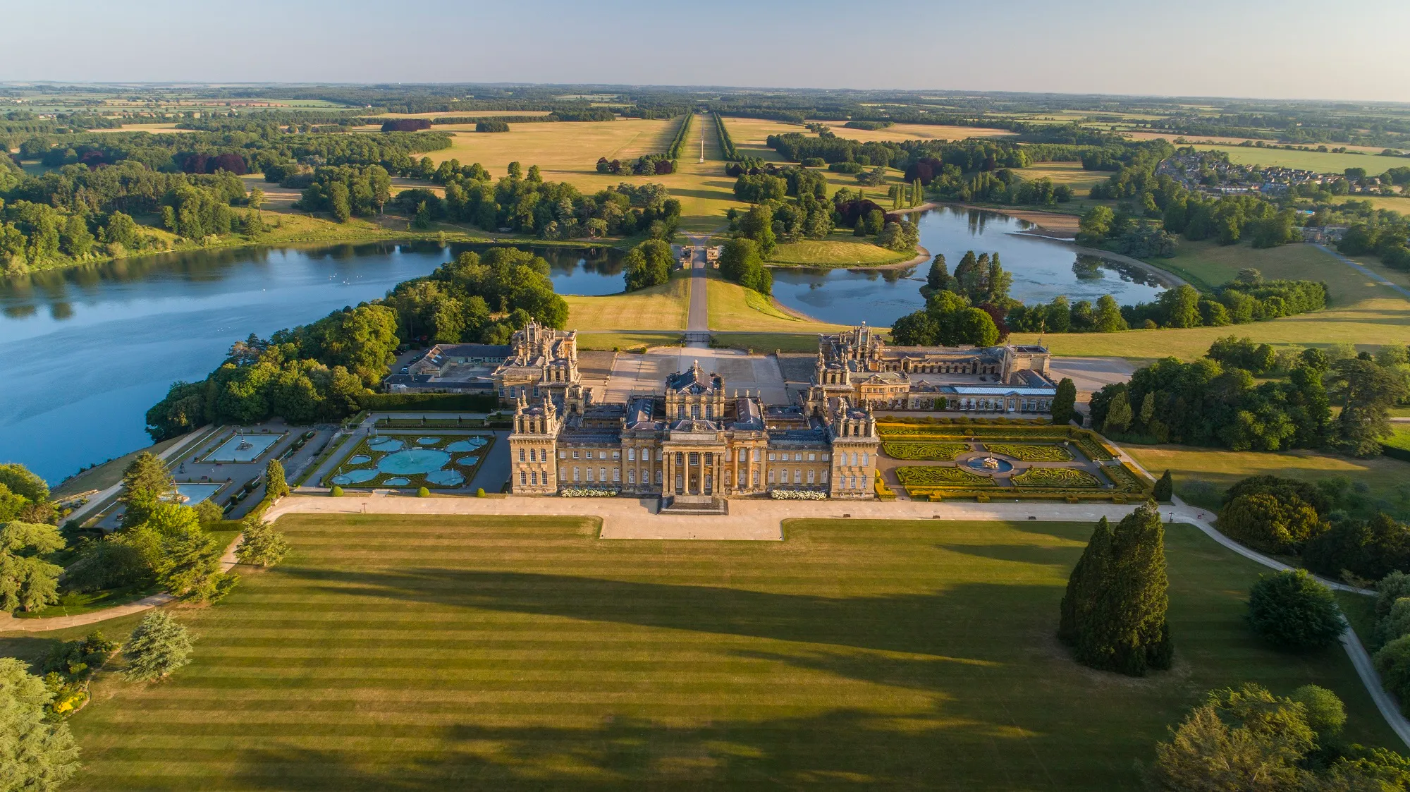 Widok z lotu ptaka na Blenheim Palace, prezentujący barokową fasadę, ogrody w stylu formalnym, jezioro Great Lake oraz otaczający teren parku podczas złotej godziny.