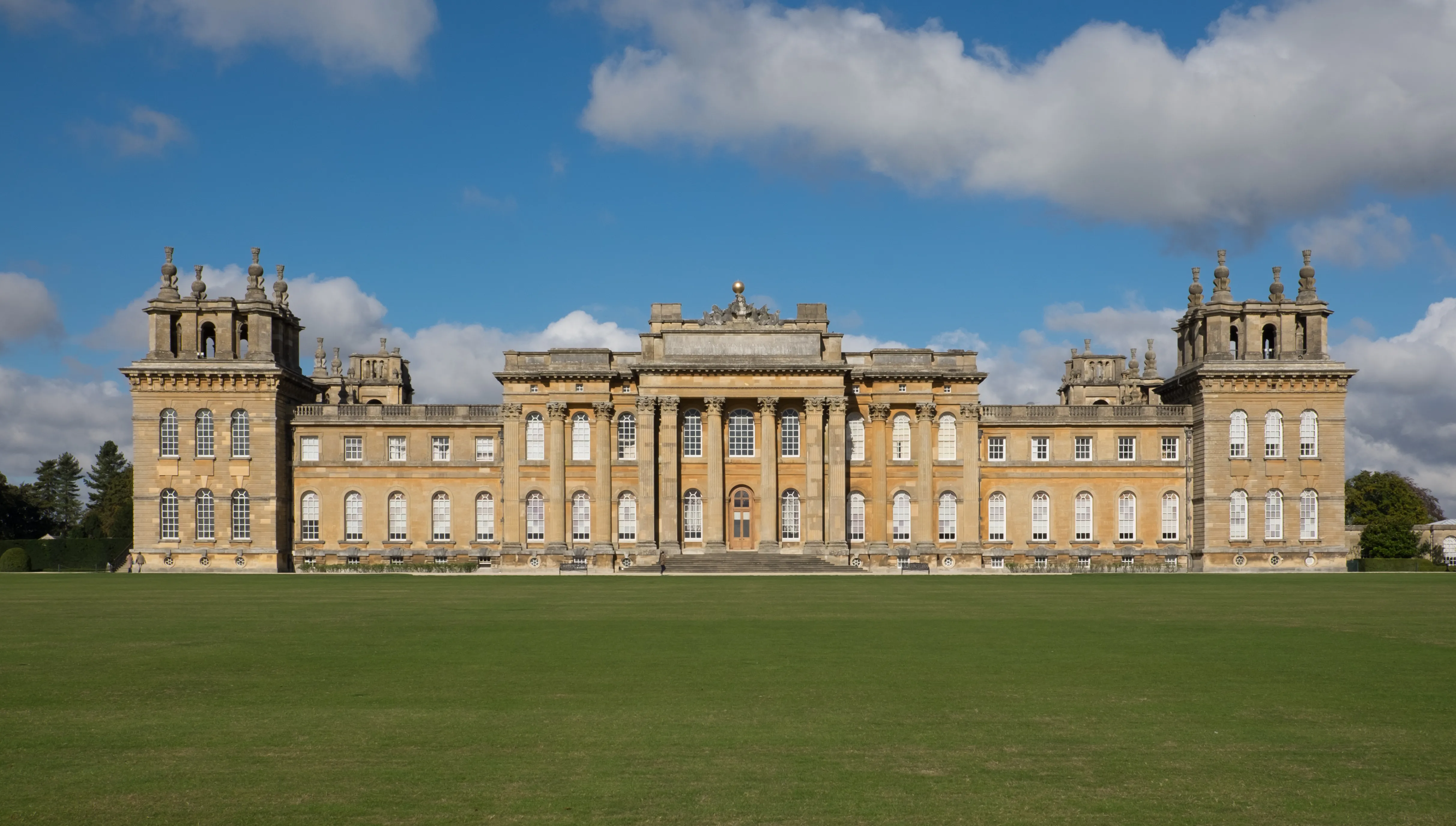 Szeroki widok na barokową fasadę Blenheim Palace z perspektywy rozległego zielonego trawnika pod niebieskim niebem z białymi chmurami