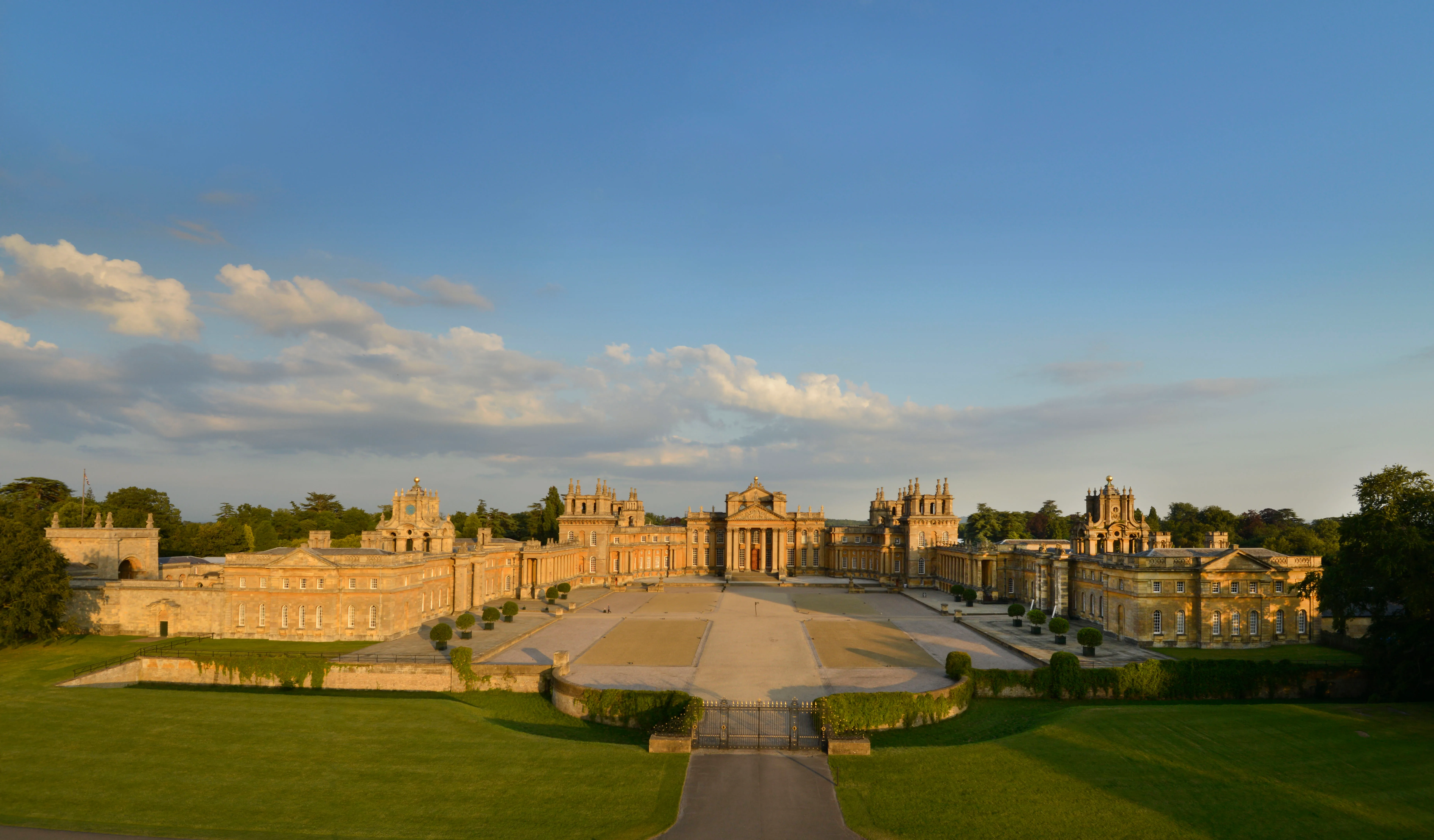 Szeroki panoramiczny widok na okazałą barokową fasadę Blenheim Palace, z symetrycznymi skrzydłami, centralnym dziedzińcem i zadbanymi trawnikami pod błękitnym niebem z rozproszonymi chmurami.
