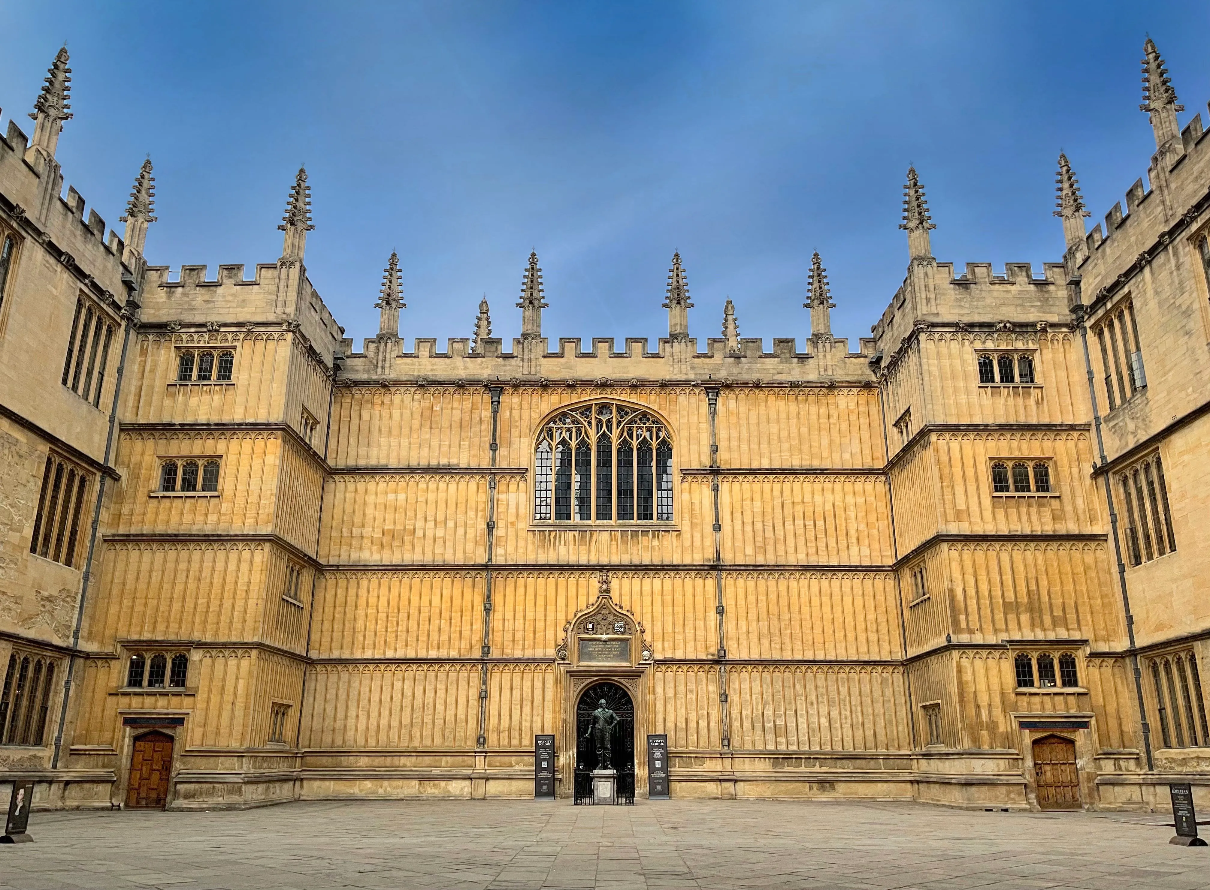 Plano general de la histórica fachada de piedra de la Bodleian Library, con sus pináculos góticos y un ventanal central arqueado bajo un cielo azul.