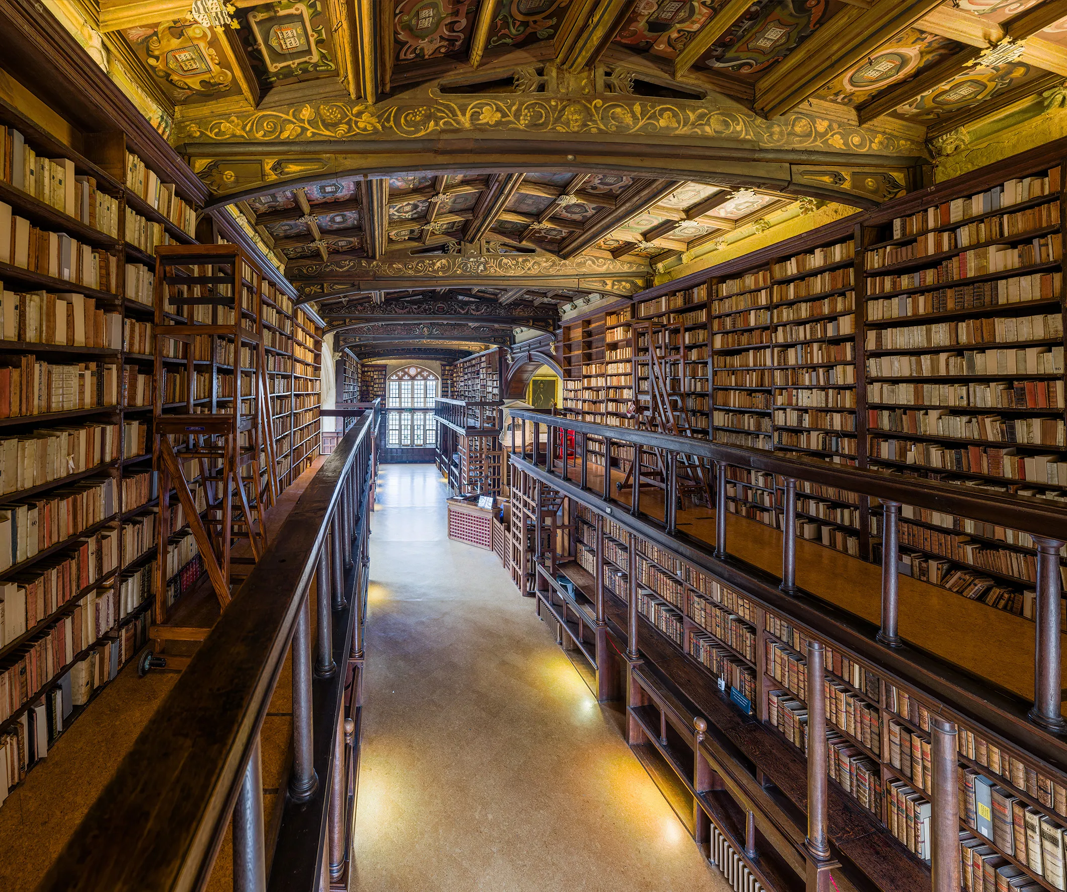 Largo pasillo de la Duke Humfrey's Library con estanterías de madera de suelo a techo llenas de libros antiguos y un ornamentado techo pintado y dorado