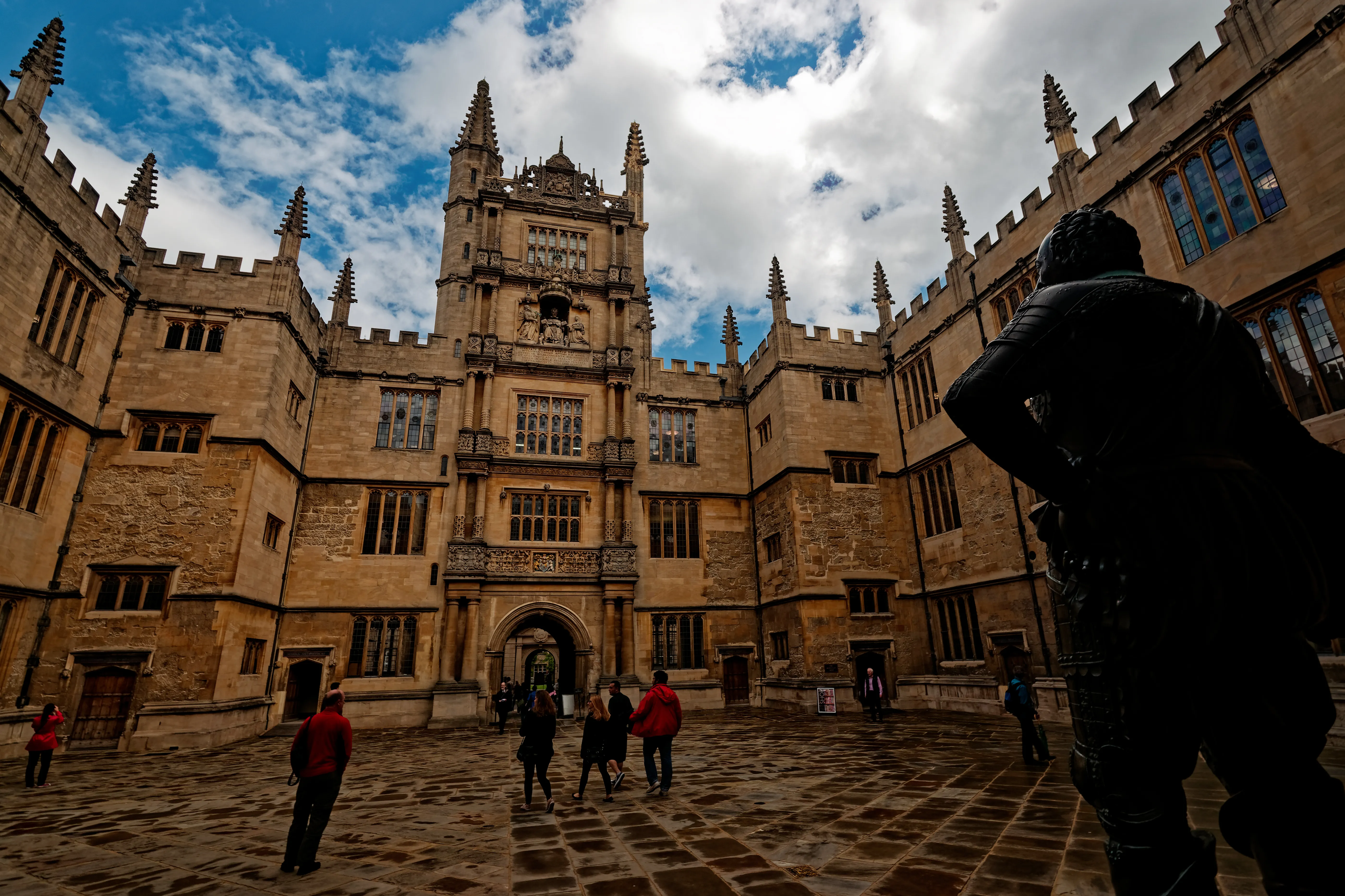 El Old School Quadrangle en la Bodleian Library, con la Tower of the Five Orders of Architecture bajo un cielo azul con nubes dispersas, y visitantes caminando por el patio empedrado.