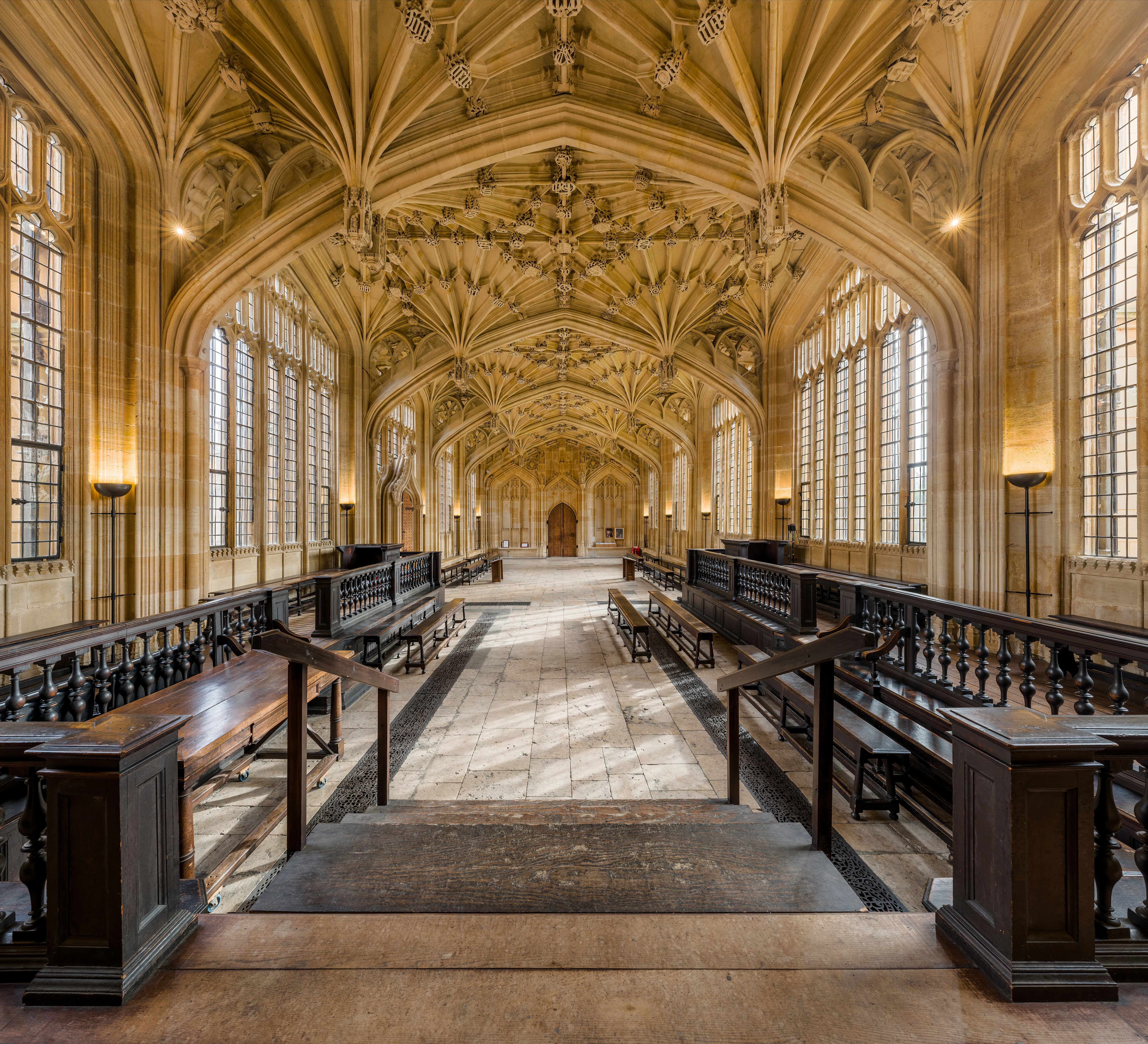 Salón largo con techo de piedra ornamentado con bóveda de abanico, ventanales con cristales emplomados y filas de bancos de madera oscura en la Divinity School de la Bodleian Library