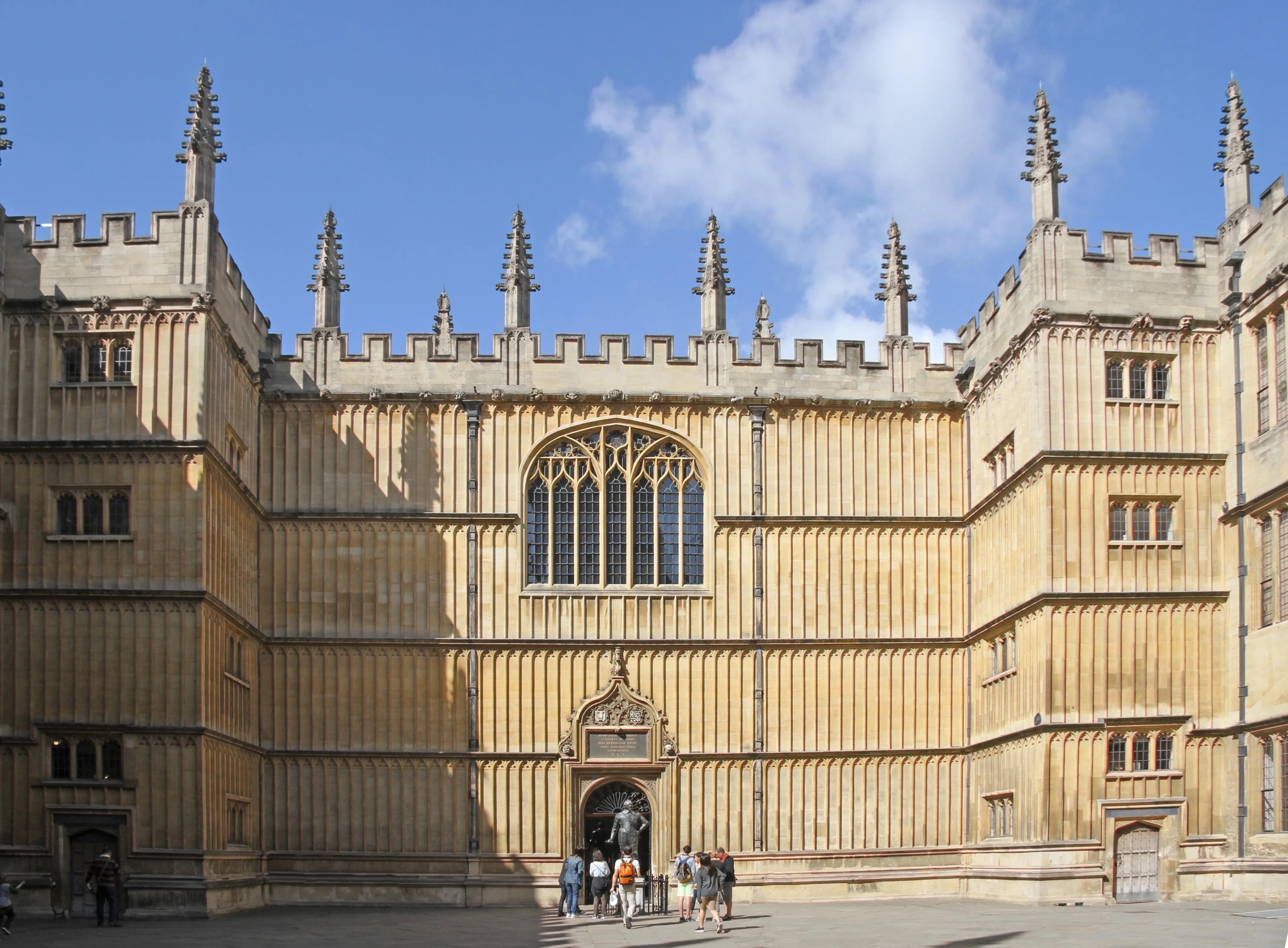 Fachada de piedra de la Bodleian Library con pináculos góticos, un gran ventanal arqueado y visitantes reunidos en la entrada central bajo un cielo azul