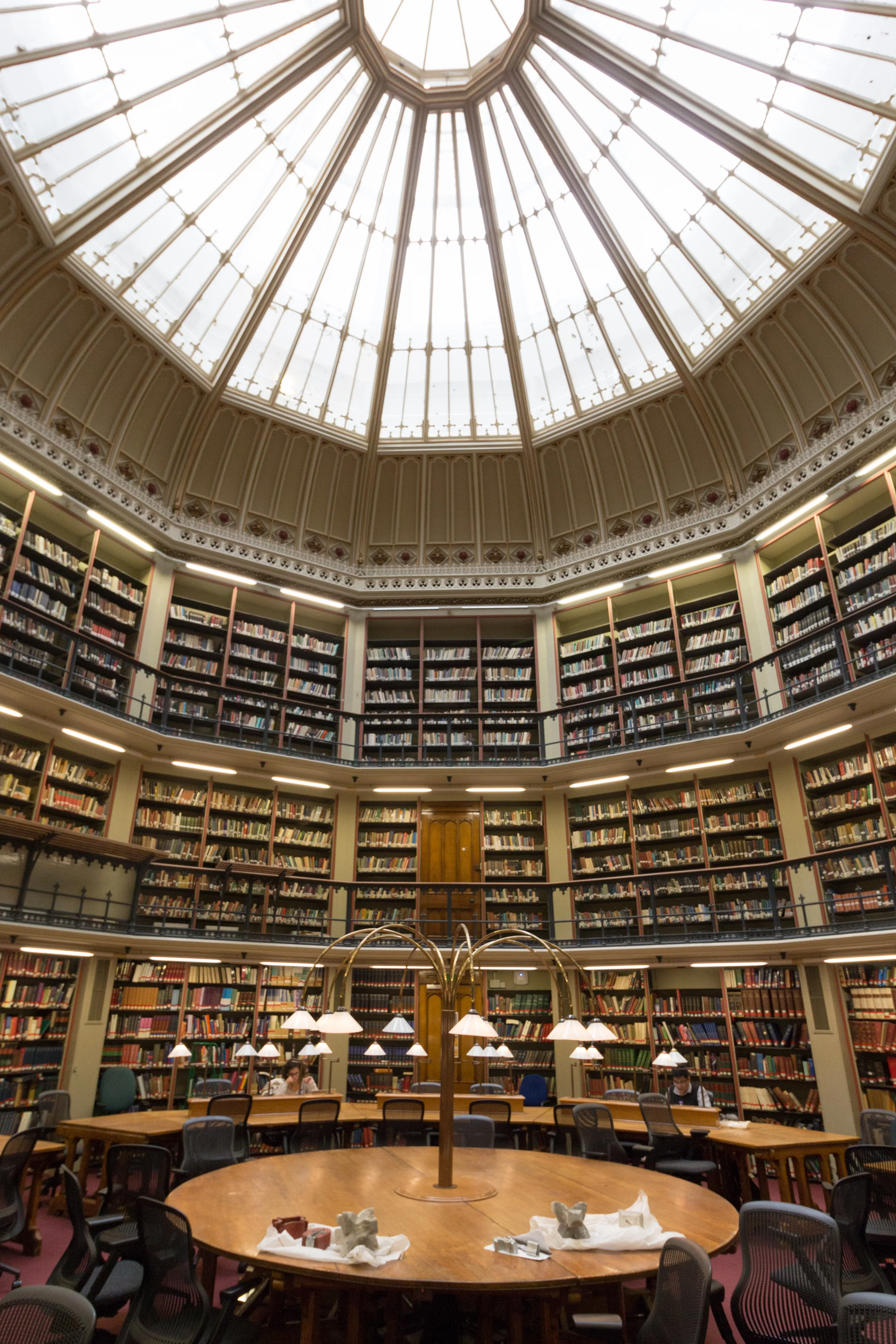 Interior de una gran sala de lectura circular de biblioteca con un gran tragaluz de cúpula de cristal, estanterías de varios niveles a lo largo de las paredes y mesas de lectura de madera con lámparas