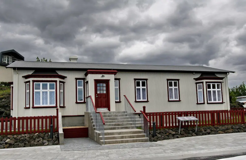 Biały parterowy dom z czerwonymi wykończeniami, czerwonym drewnianym płotem i betonowymi schodami prowadzącymi do czerwonych drzwi wejściowych pod zachmurzonym niebem
