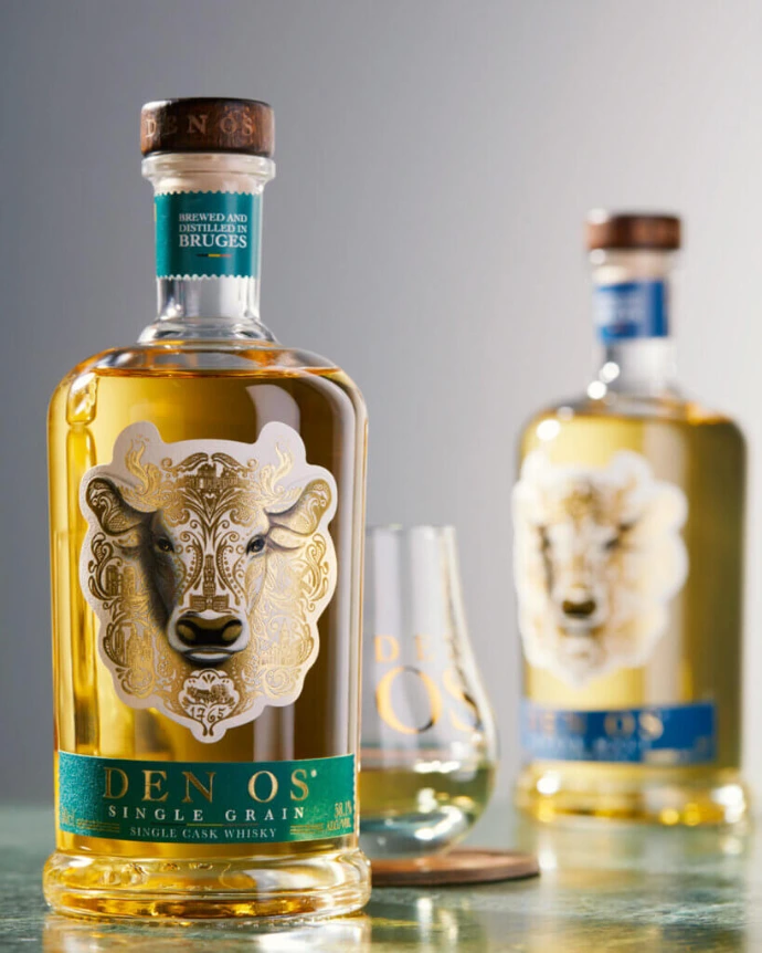 Dos botellas de Den Os Single Grain Belgian Grain Whisky con líquido color ámbar y etiquetas ornamentadas con cabeza de toro, flanqueando una copa de degustación en forma de tulipán sobre una superficie reflectante.