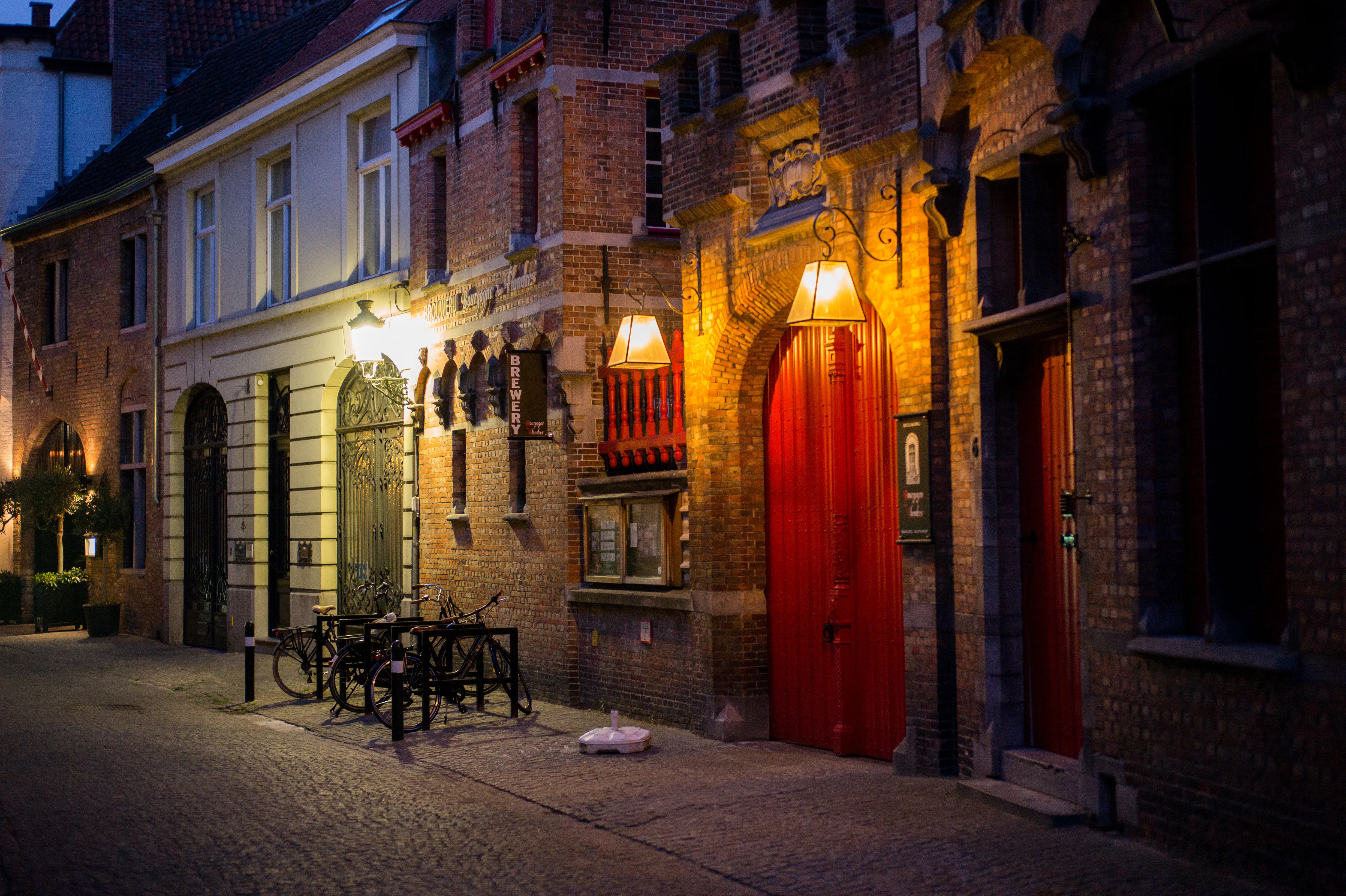 Vista nocturna de la calle con edificios históricos de ladrillo, una puerta roja iluminada y bicicletas aparcadas sobre un camino de adoquines.