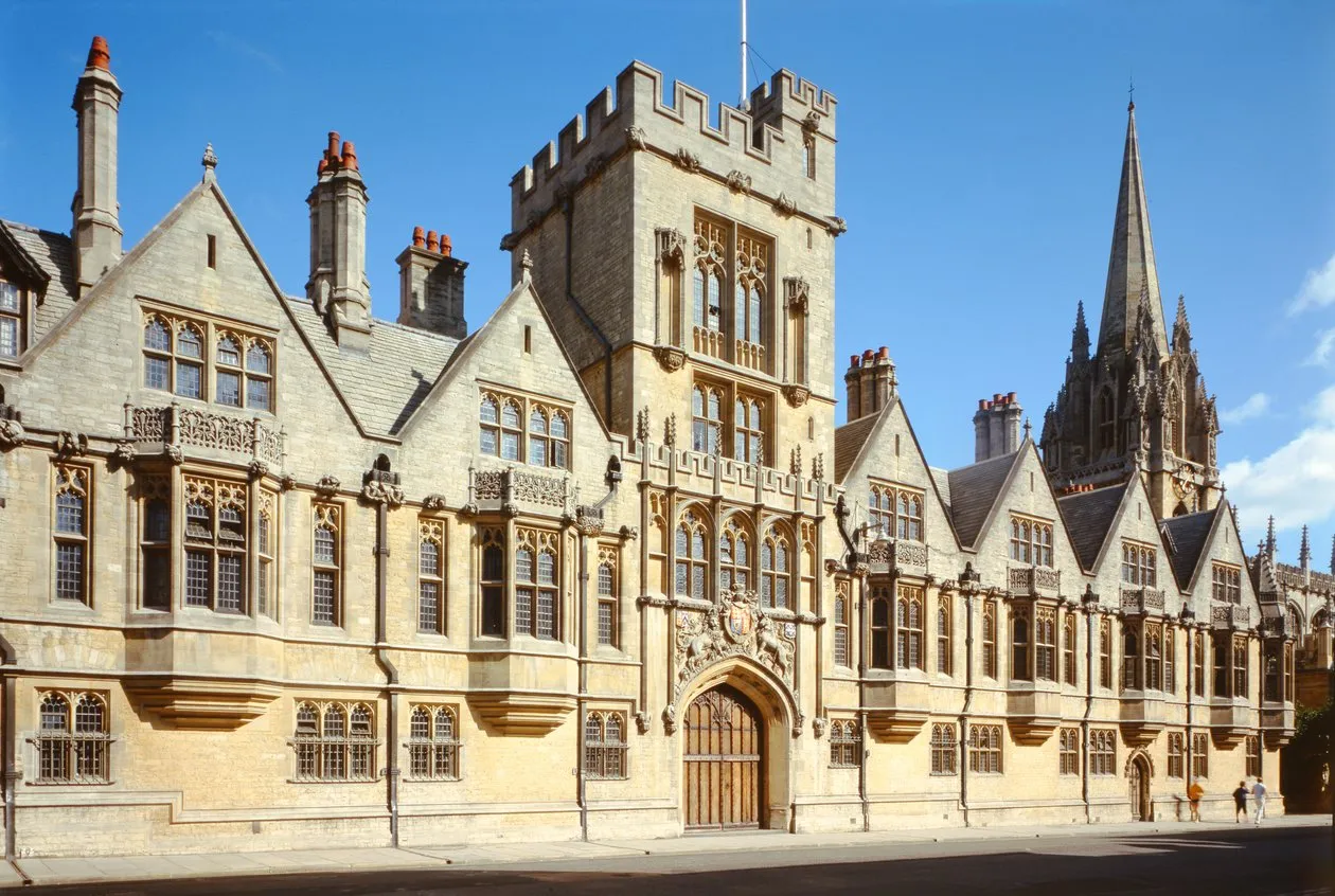 Widok z zewnątrz na zabytkową kamienną fasadę Brasenose College z gotycką wieżą, ozdobnymi oknami dzielonymi słupkami, wieloma kominami oraz iglicą All Saints Church w tle pod błękitnym niebem.