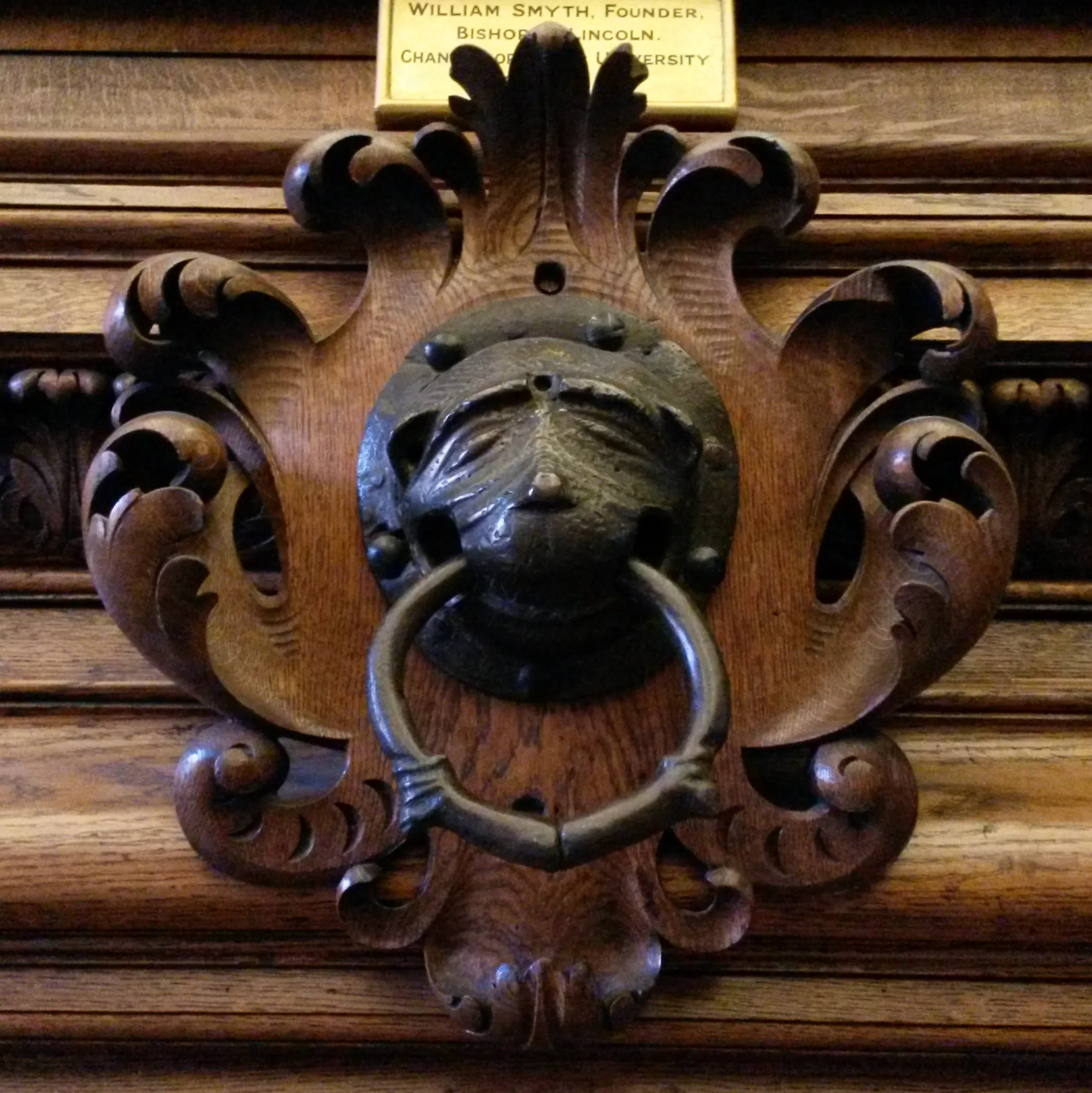 Ozdobna brązowa kołatka w kształcie twarzy z pierścieniem, zamontowana na rzeźbionym drewnianym panelu z małą tabliczką powyżej o treści 'William Smyth, Founder Bishop of Lincoln Chancellor University'.