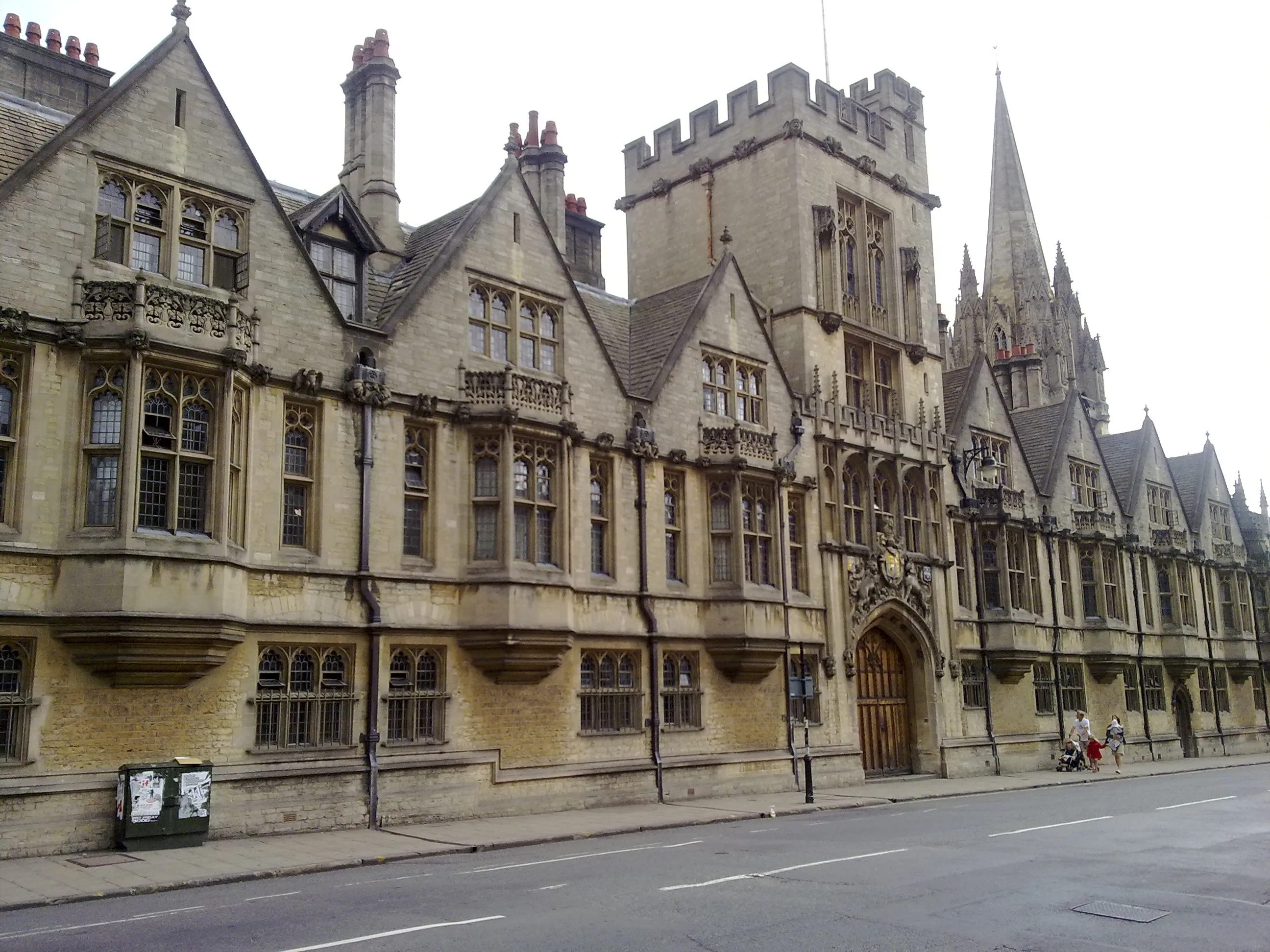Historyczna kamienna fasada Brasenose College wzdłuż High Street, z gotyckimi szczytami, centralną wieżą z krenelażem, łukową drewnianą bramą oraz iglicą kościoła w tle.