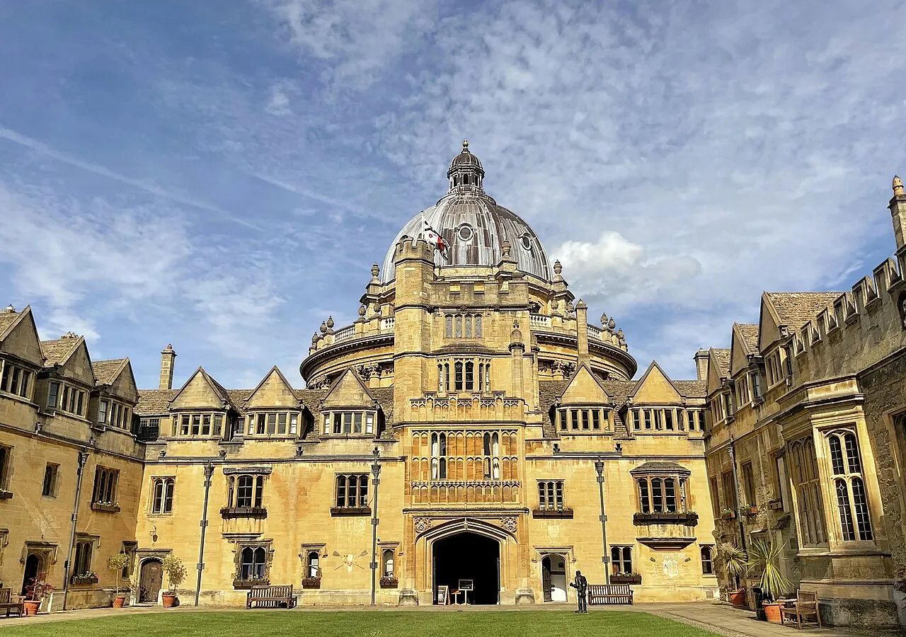Duży budynek Radcliffe Camera z kopułą widziany z dziedzińca college'u, w otoczeniu zabytkowych kamiennych gmachów, zielonego trawnika oraz błękitnego nieba z rozproszonymi chmurami.