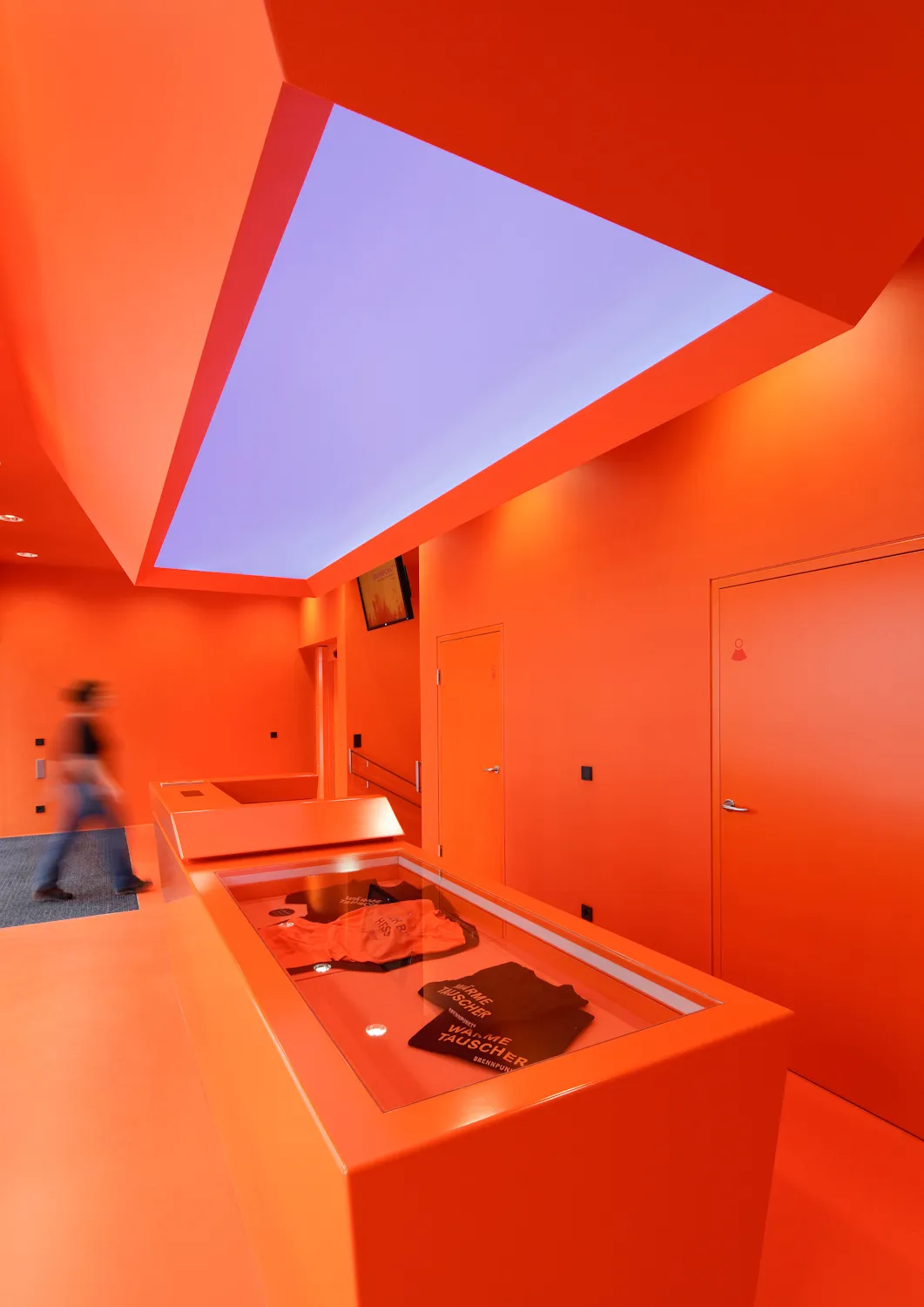 Interior de un museo con paredes y techo de color naranja brillante, una vitrina de cristal y una persona caminando al fondo.