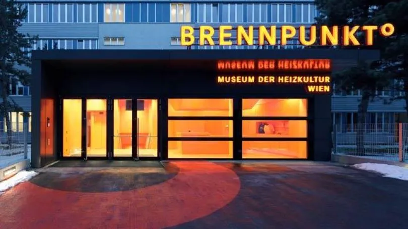 La entrada iluminada del Museo Brennpunkt de Cultura de la Calefacción por la noche, con luz naranja brillando a través de las puertas de vidrio y el nombre mostrado en letreros de neón.
