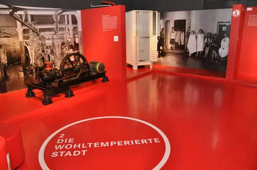 Sala de exposiciones con suelo y paredes rojos, una gran máquina industrial y fotografías históricas en exhibición.