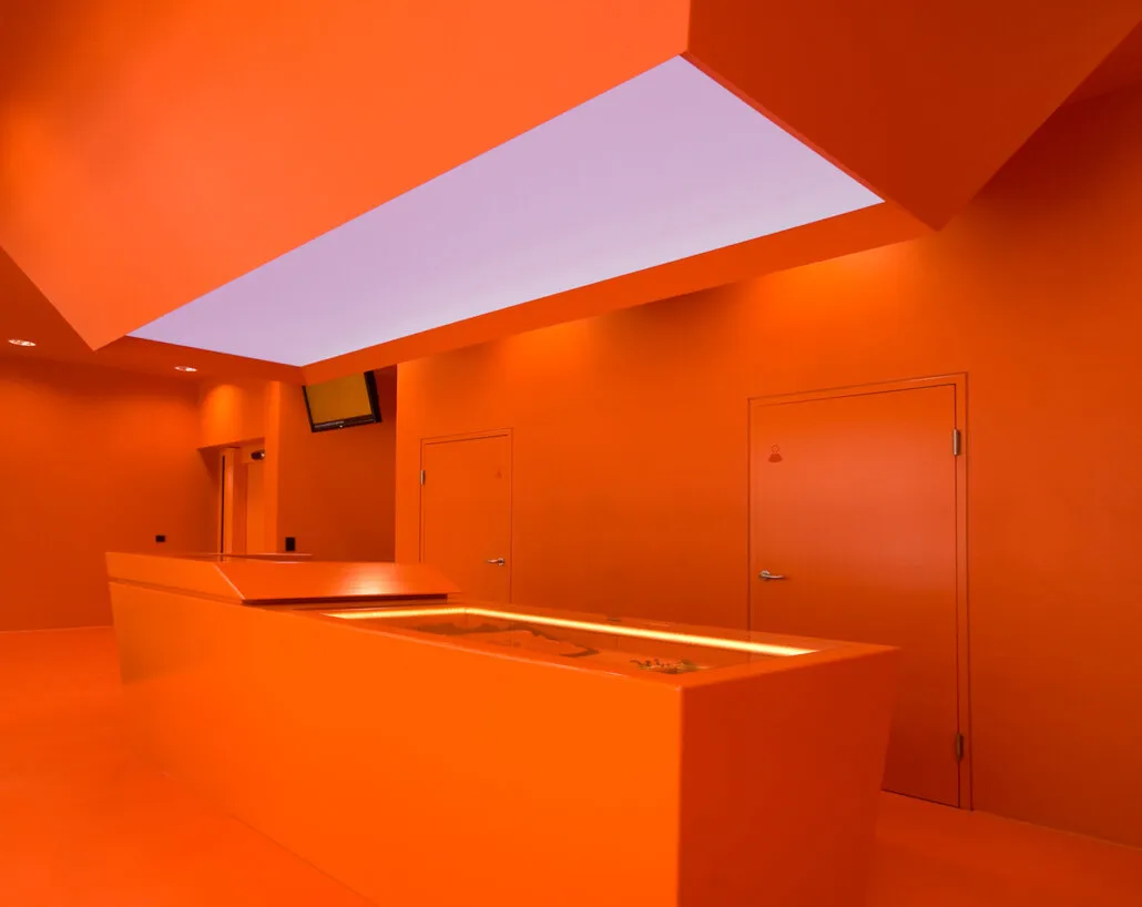 Interior de un museo con paredes naranjas y un mostrador central bajo un tragaluz