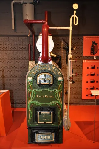 Una estufa de calefacción vintage verde y negra con tuberías rojas y blancas, etiquetada como 'Rapid Ketel', expuesta en una galería de museo con suelo naranja y un panel de pared rojo.