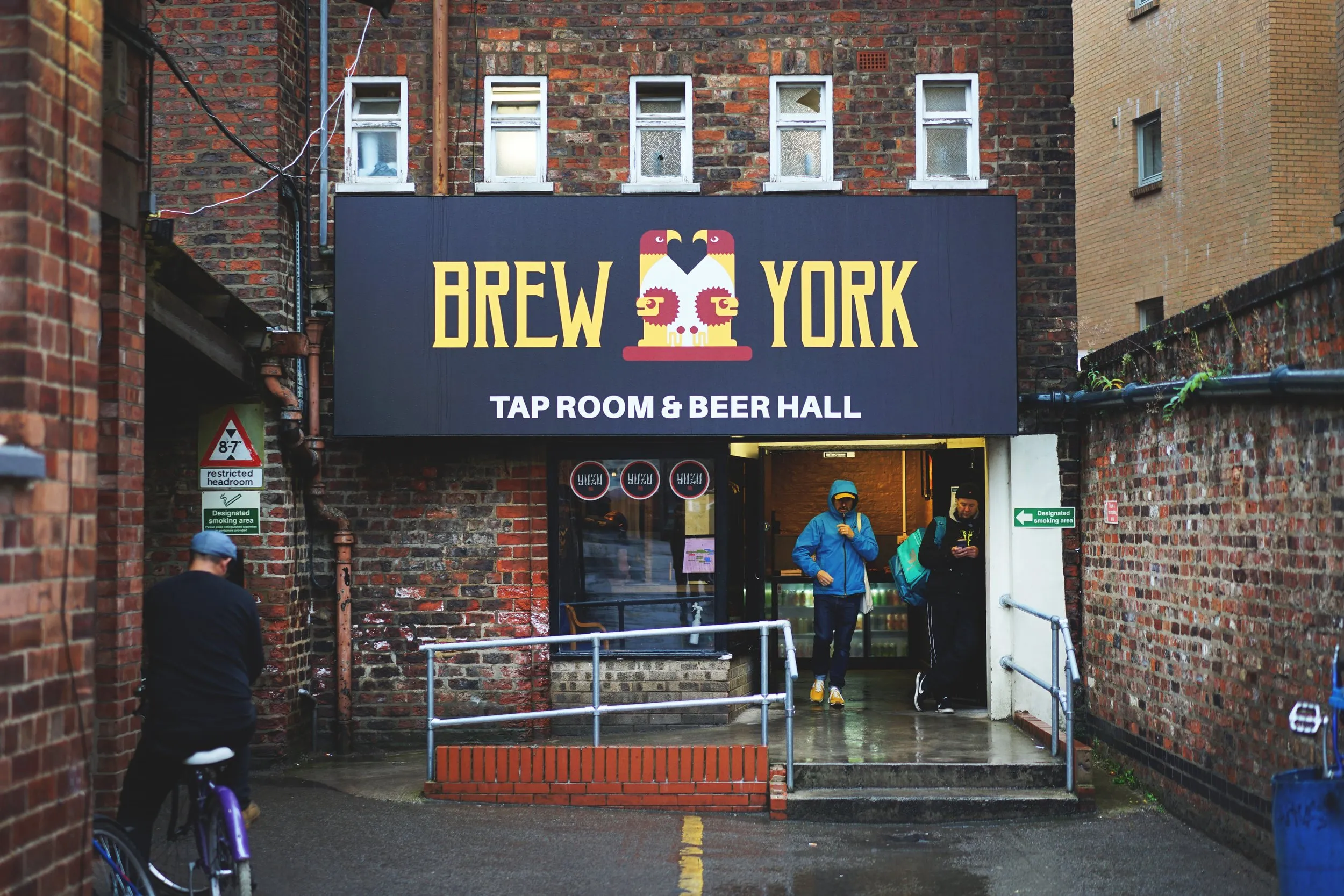 Widok z zewnątrz na Brew York Tap Room & Beer Hall z dużym czarnym szyldem nad wejściem, ceglaną elewacją budynku i ludźmi stojącymi przy drzwiach