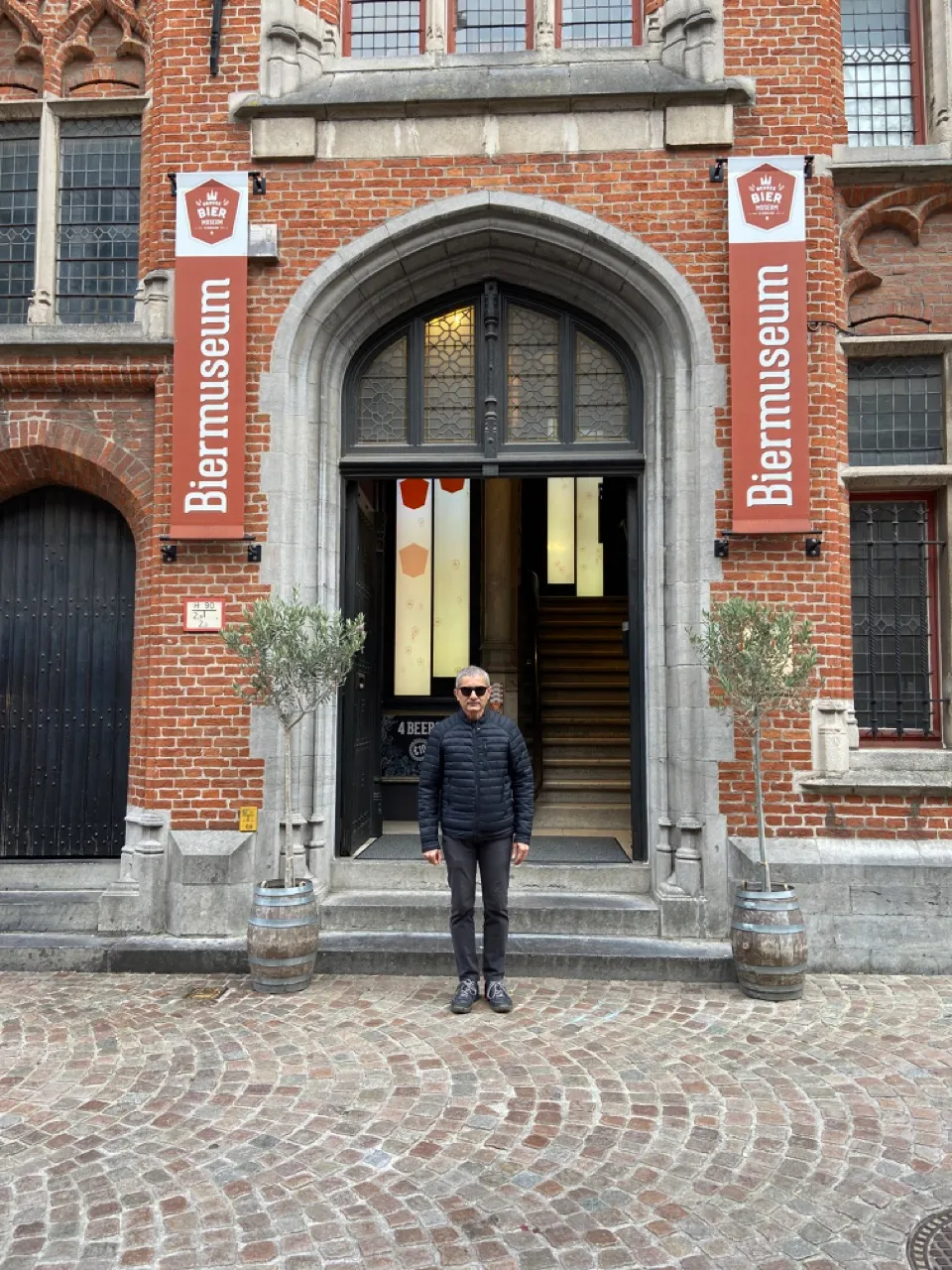 Een man staat op een kasseienstraat voor de ingang van een bakstenen gebouw, gemarkeerd door twee verticale rode banieren met de tekst 'Biermuseum'.