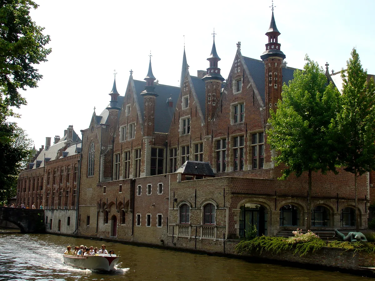 Gran edificio histórico de ladrillo rojo con varias agujas puntiagudas a lo largo de un canal en Brujas, con un barco turístico pasando por el primer plano.