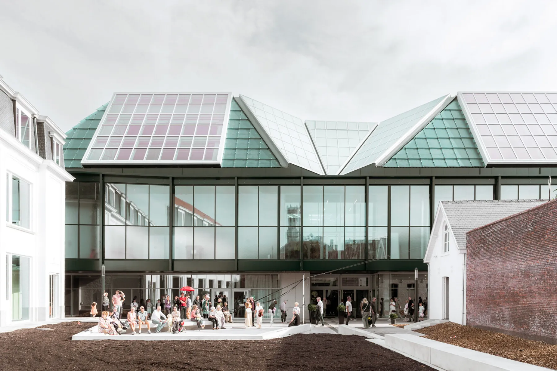 Edificio moderno con distintivos paneles de techo de cristal verde en ángulo y paneles solares, gran fachada de cristal, con personas reunidas en el exterior sobre una zona pavimentada
