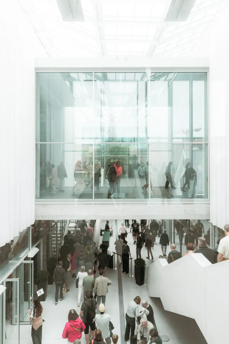 Vista en ángulo elevado del interior de una galería moderna que cuenta con una pasarela superior acristalada con vistas a un atrio de paredes blancas lleno de visitantes.