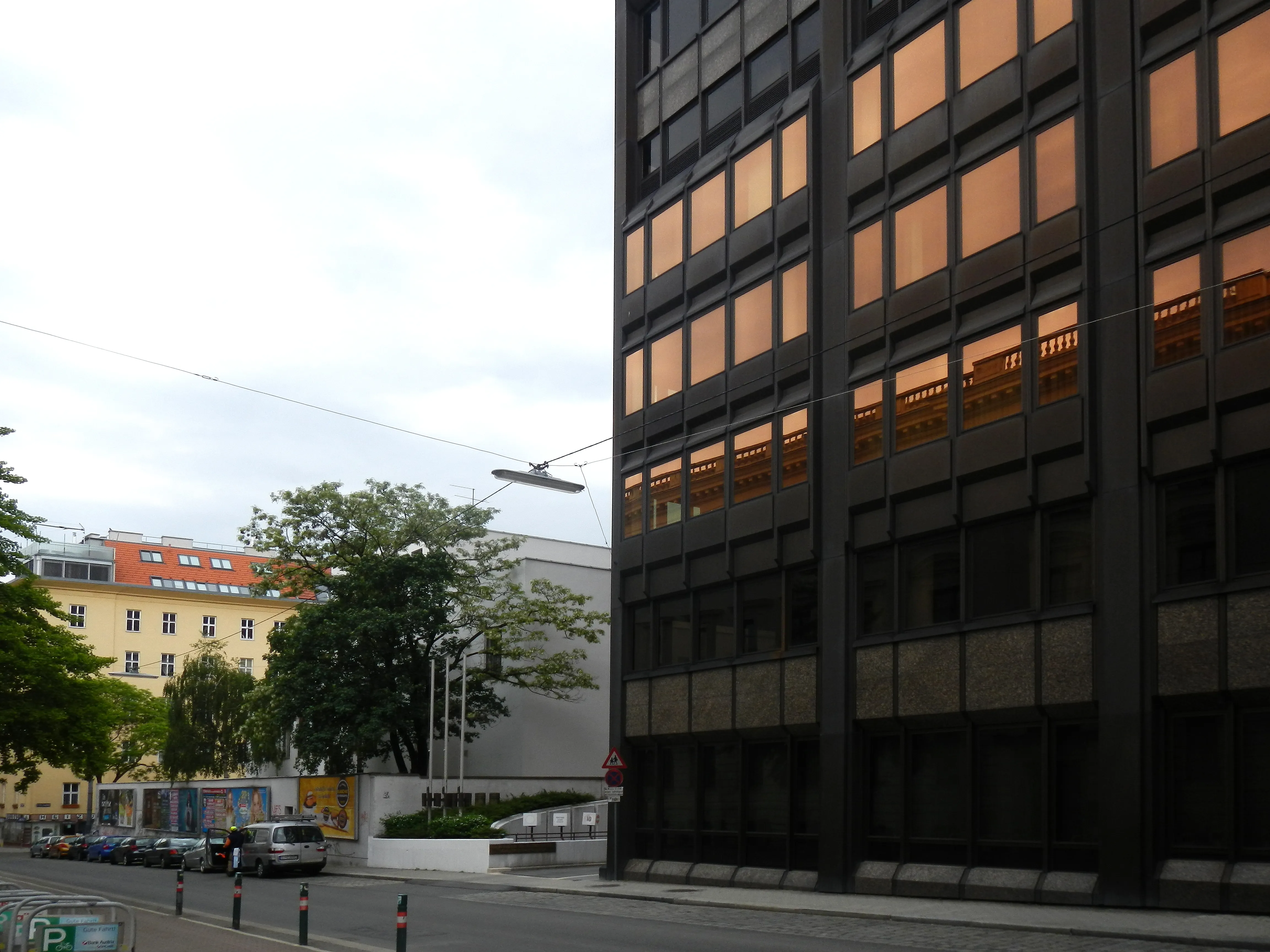 Edificio modernista con fachada oscura y grandes ventanales, vista a la calle con árboles y otros edificios al fondo