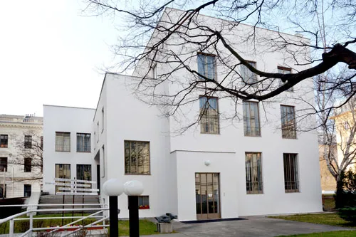 Edificio modernista blanco con ventanas rectangulares y una fachada minimalista, rodeado de ramas de árboles desnudas y una pequeña zona ajardinada.