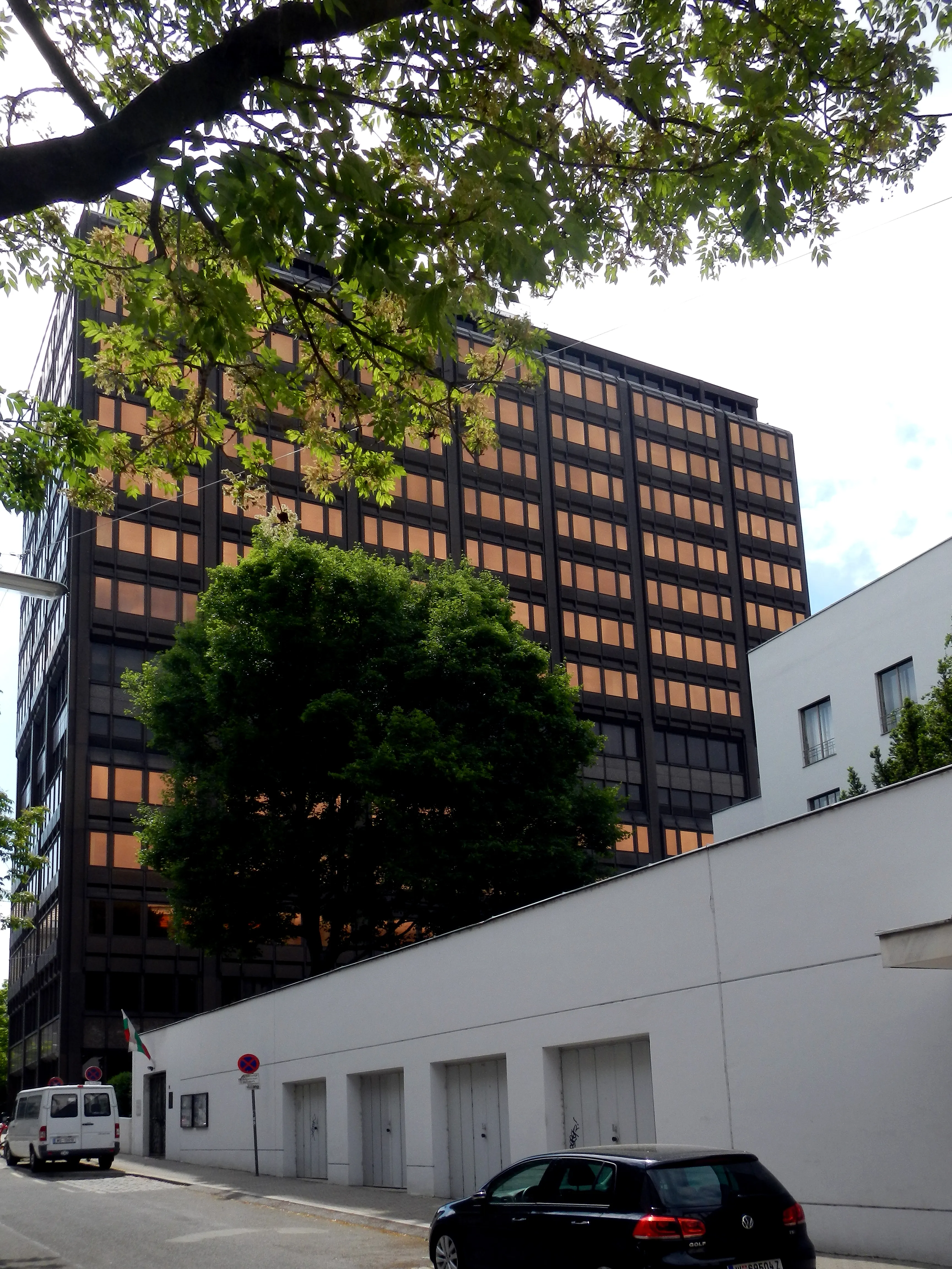 Un edificio modernista con una cuadrícula de ventanas iluminadas, parcialmente oscurecido por árboles y un muro blanco