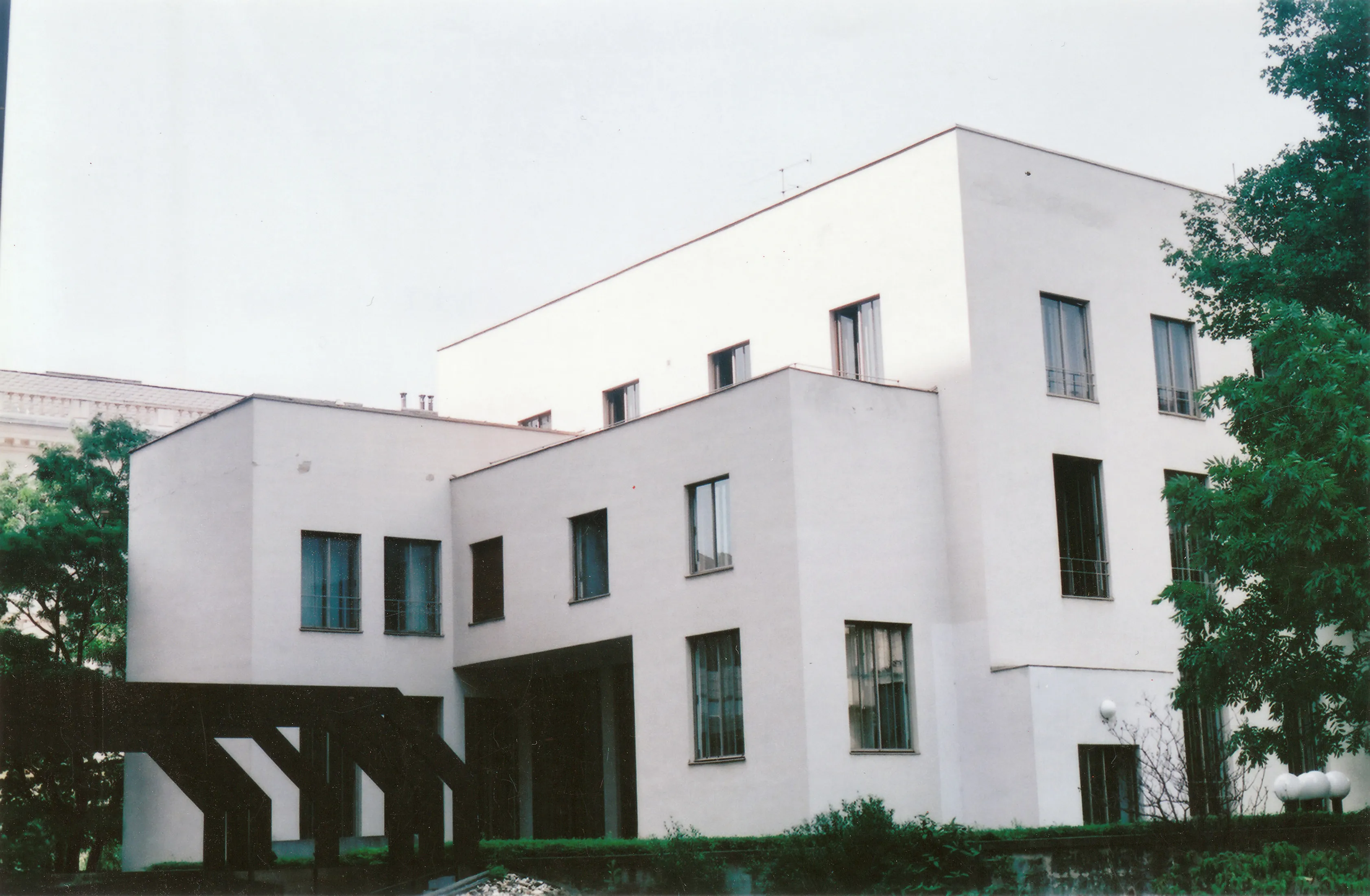 Edificio modernista blanco con diseño geométrico y grandes ventanales, rodeado de árboles y vegetación.