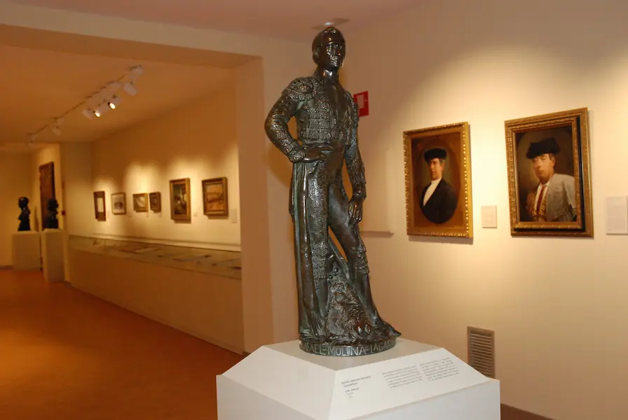 Bronzen beeld van een stierenvechter in een museumgalerij met ingelijste portretten aan de muur