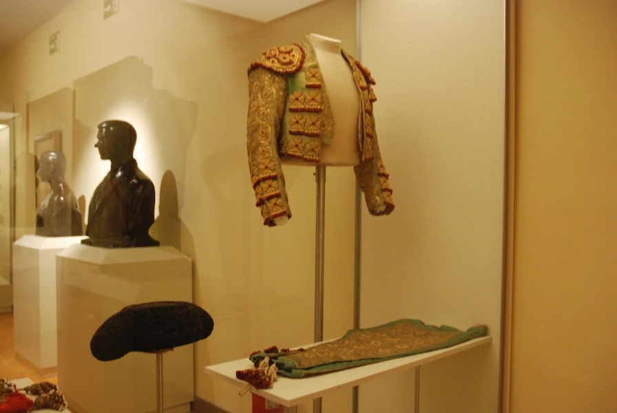 Een traditioneel torero-jasje aan een paspop, een bustesculptuur en een hoed tentoongesteld in een museumgalerij.