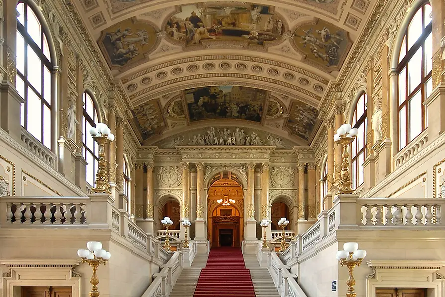 Sierlijk interieur van het Burgtheater met een rood beklede trap, gouden balustrades en een plafond versierd met fresco's en architectonische details