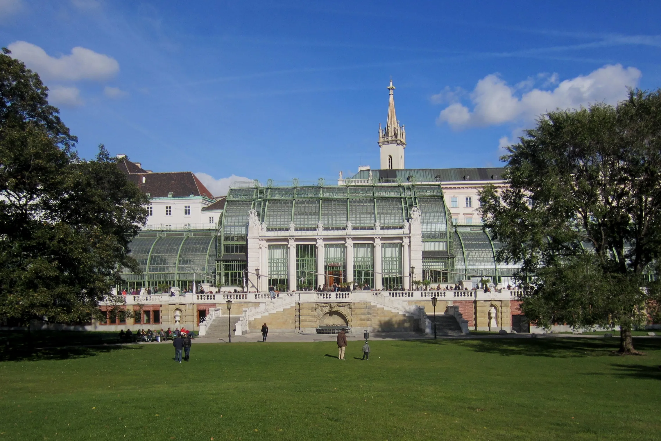 Palmiarnia w Burggarten z szklaną oranżerią i centralnymi schodami, otoczona zielonymi trawnikami i drzewami pod błękitnym niebem.