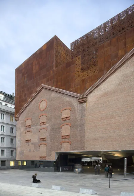 Buitenkant van CaixaForum Madrid, een gebouw met een bakstenen gevel en een grote roestbruine metalen structuur met een verticale tuin, met mensen die ervoor lopen.