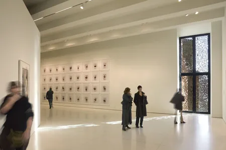 Grote witte galerieruimte met een muur van ingelijste kunstwerken, mensen die rondlopen en kijken, en een hoog decoratief raam aan de rechterkant.
