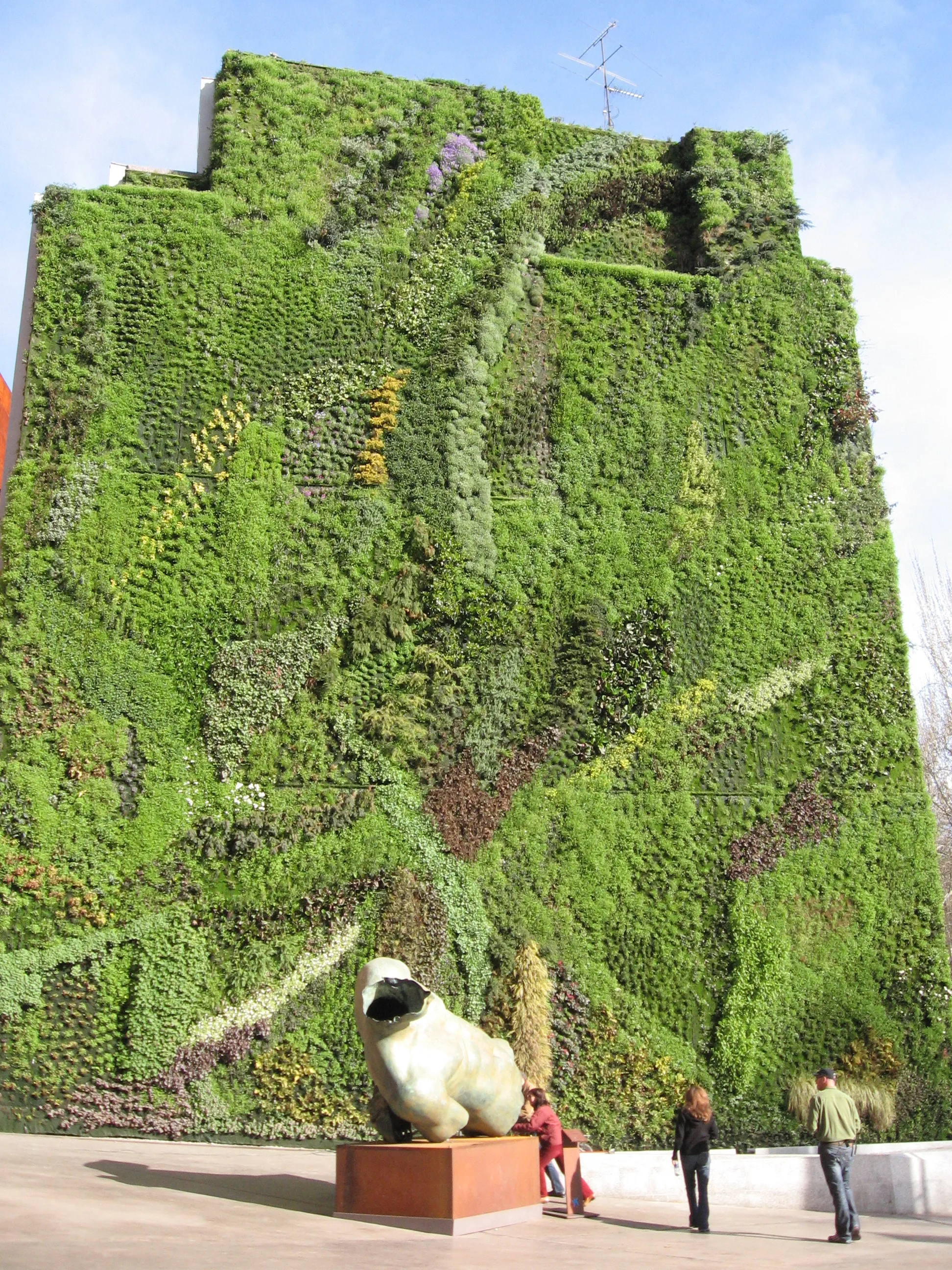 Een hoog gebouw bedekt met een dichte groene verticale tuin met een wit schaapsculptuur ervoor en mensen die op de stoep lopen