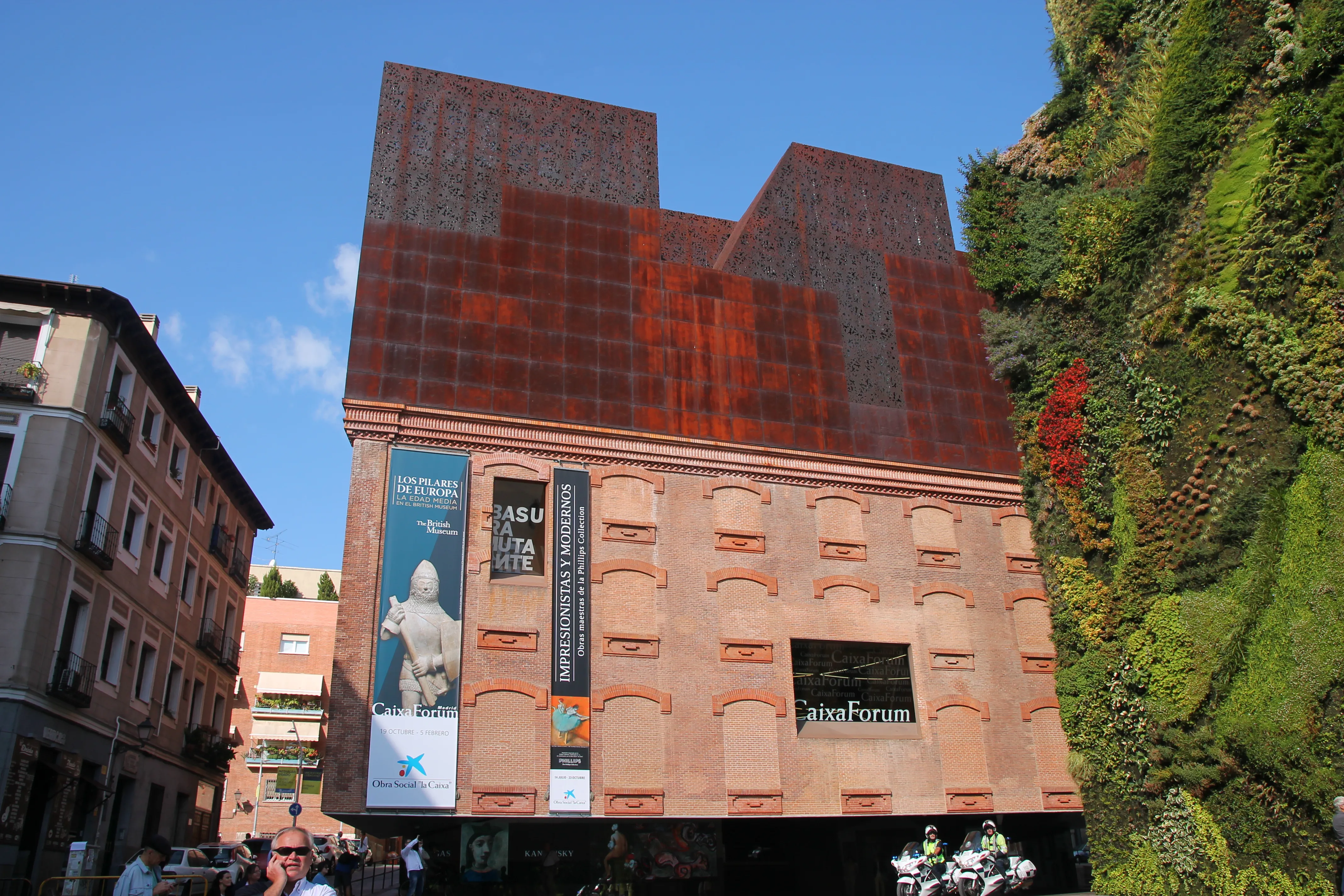 Het CaixaForum Madrid gebouw met een verticale tuin aan de rechterkant en een roestbruine metalen gevel, onder een blauwe lucht.