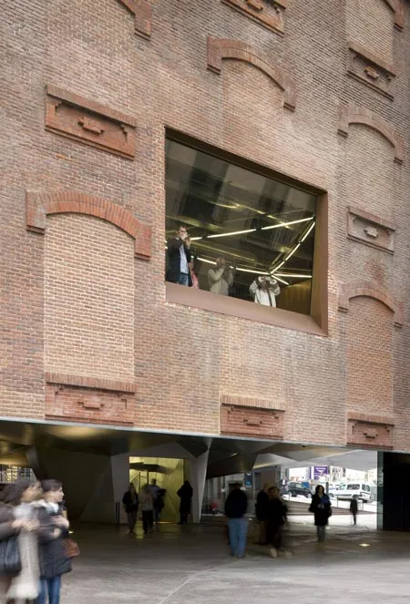 Bakstenen gevel van CaixaForum Madrid met groot raam dat interieurverlichting en mensen binnen toont