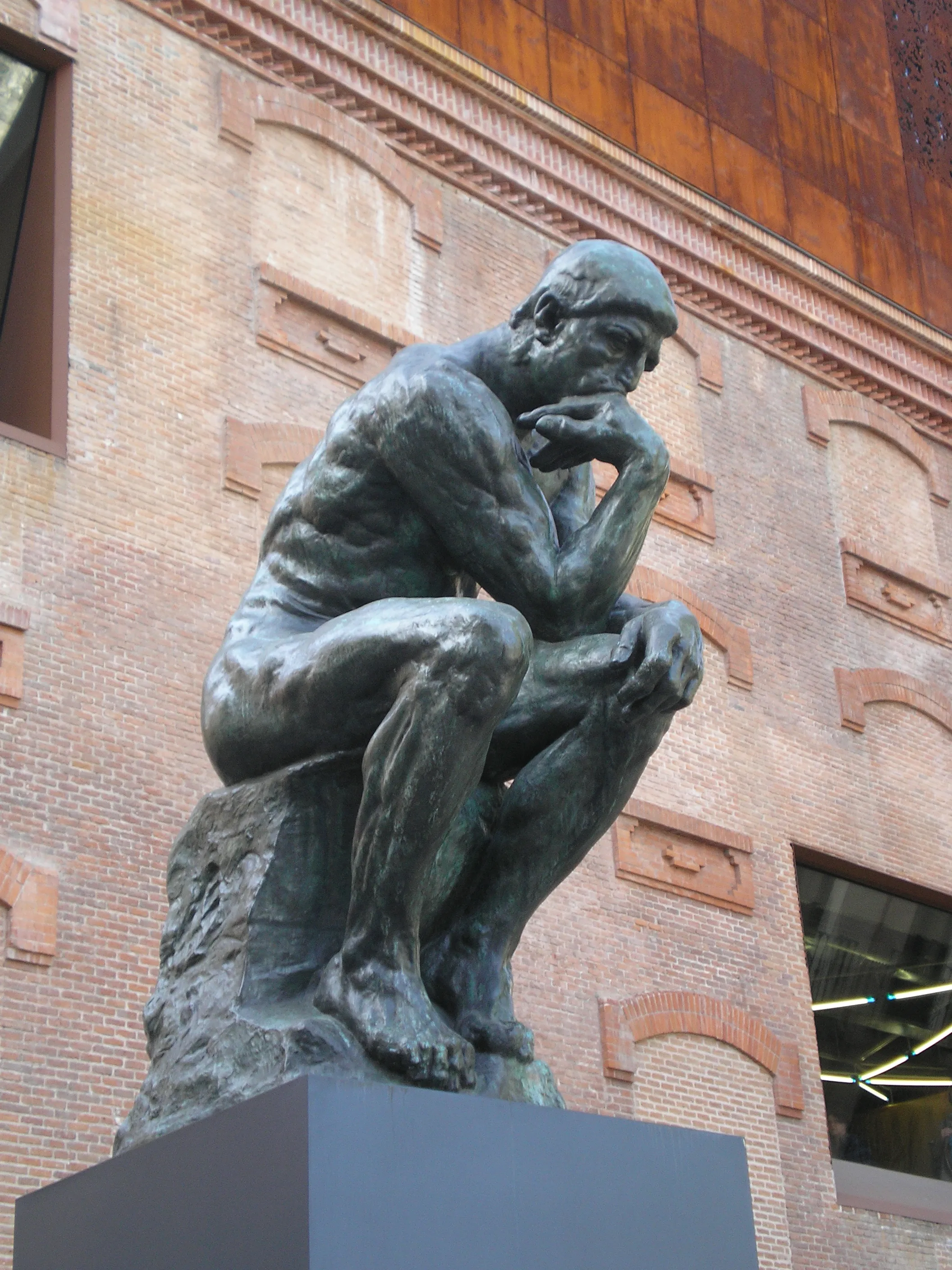 Bronzen beeld van een zittende man met de kin op de hand, tegen een bakstenen muur met rondboogvensters