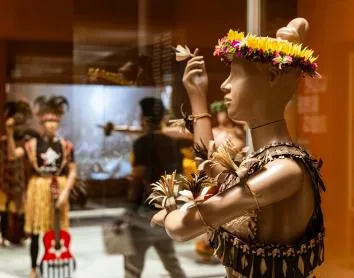 Paspop met traditionele Pacifische eilandkleding, een bloemenhoofddeksel en geweven accessoires, tentoongesteld in een museumexpositie.