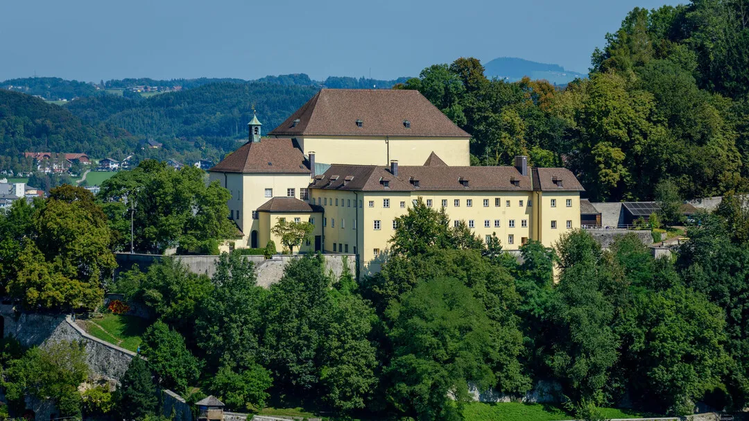 A large yellow monastery building with a brown roof and church tower sits atop a wooded hill, with green trees in the foreground and a cityscape and hills in the background.