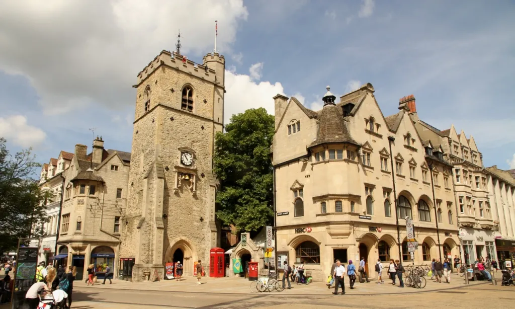 Stenen middeleeuwse toren met een wijzerplaat die boven de omliggende historische gebouwen uitsteekt op een druk kruispunt in Oxford, met voetgangers en fietsers op de voorgrond.