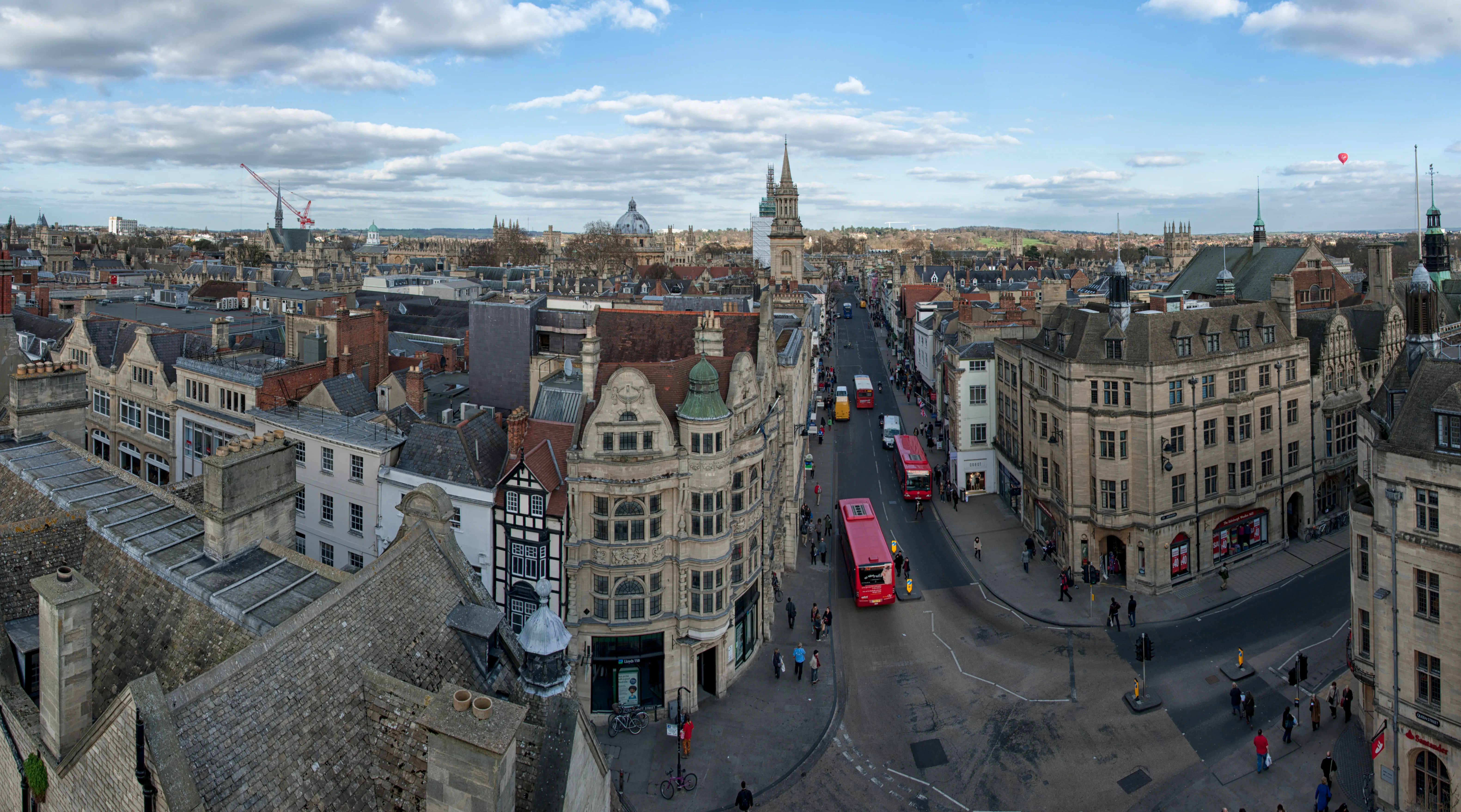 Panoramisch uitzicht over het centrum van Oxford vanaf de Carfax Tower, met historische stenen gebouwen, een straat met een rode dubbeldekkerbus en kerktorens onder een halfbewolkte lucht.