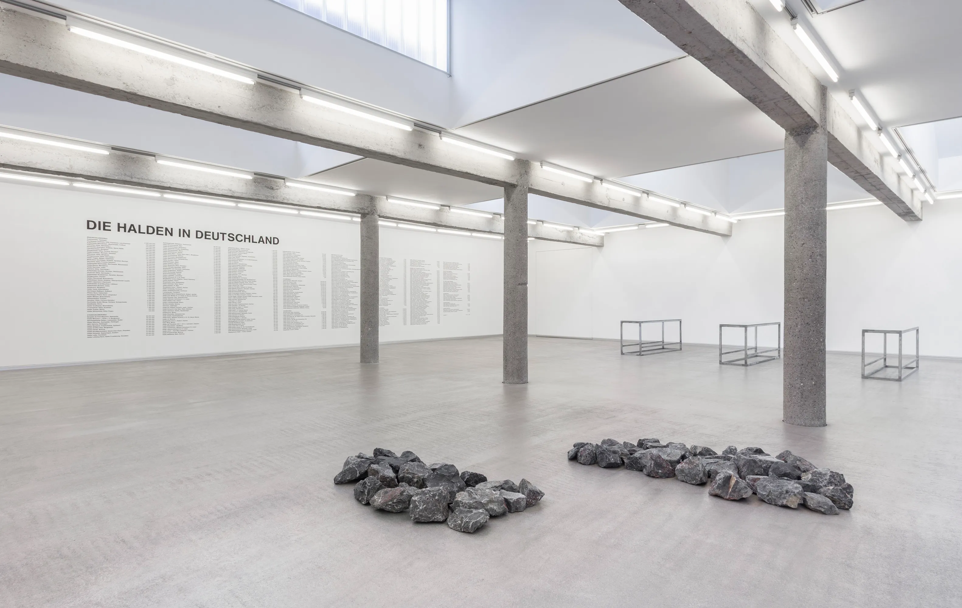 Gallery interior with wall text reading 'DIE HALDEN IN DEUTSCHLAND' and piles of dark rocks arranged on the concrete floor
