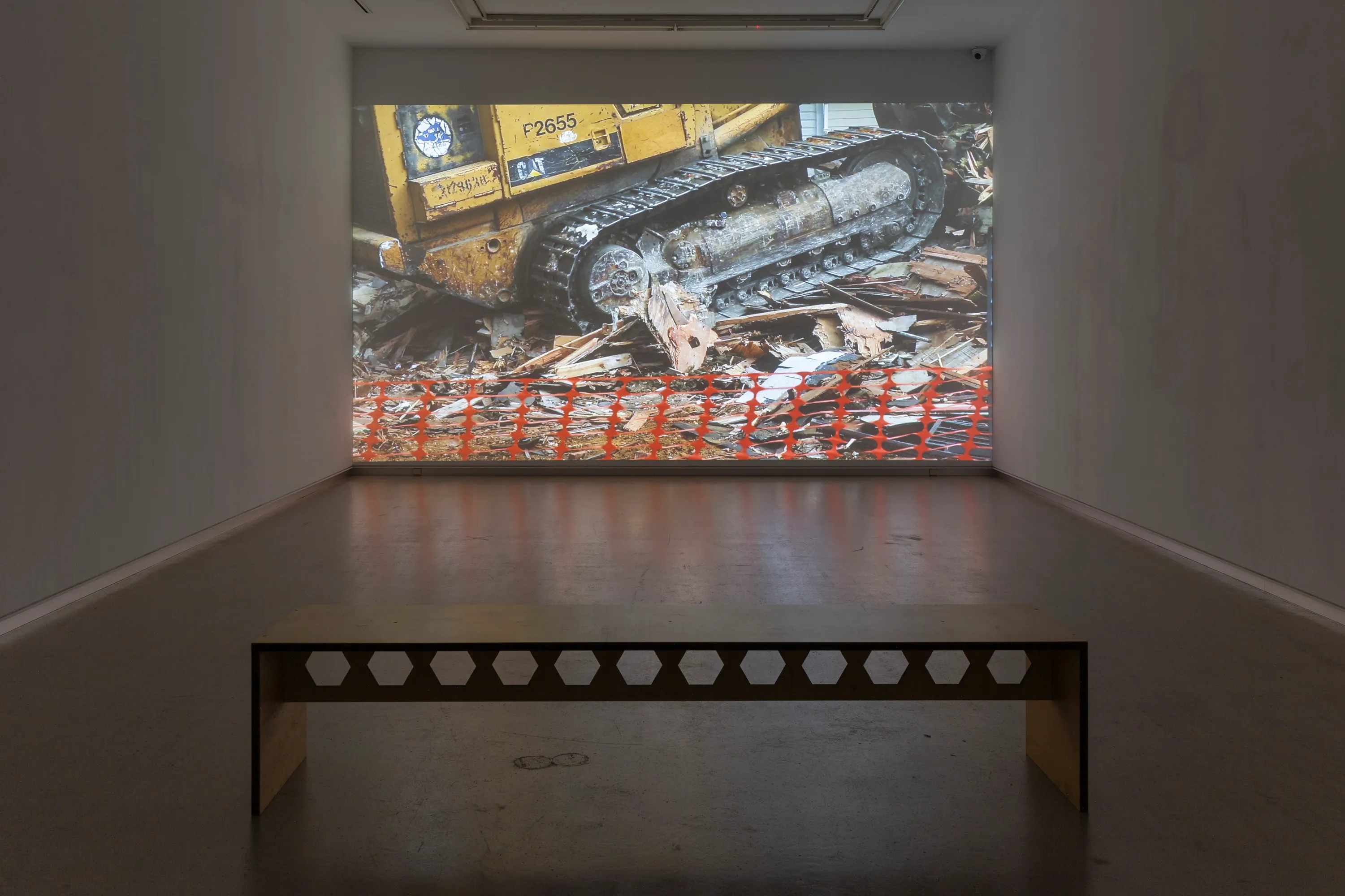 Gallery room with a large wall projection of a yellow tracked bulldozer on rubble and orange barriers, with a perforated metal bench in the foreground
